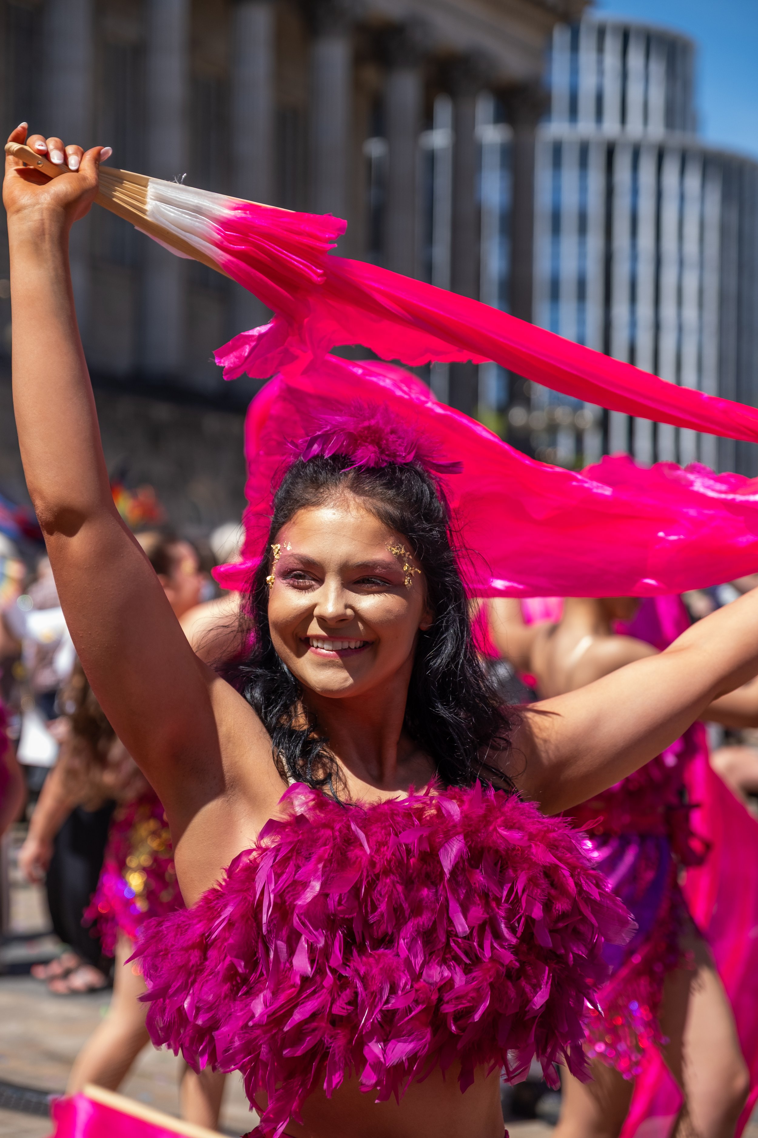 A woman smiling and dancing in a bright pink feathered costume during a parade or celebration, with a crowd and modern buildings in the background.