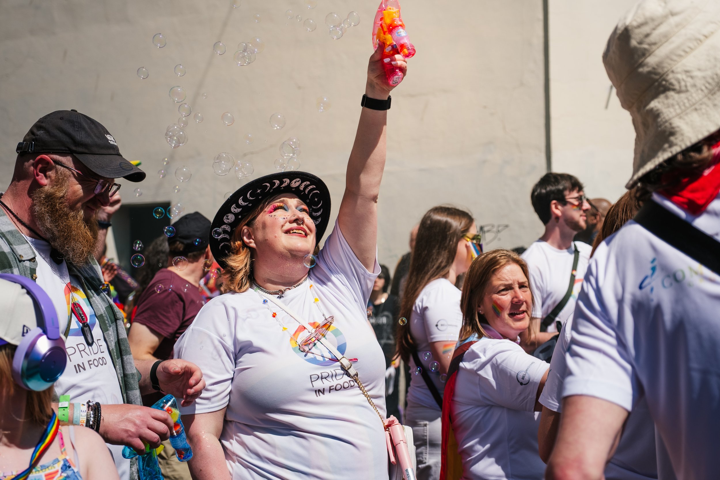 A group of diverse people participating in a Pride event outdoors. A woman with a hat and colorful makeup is holding bubbles and smiling. Others around her are also smiling, wearing Pride-themed clothing and accessories.