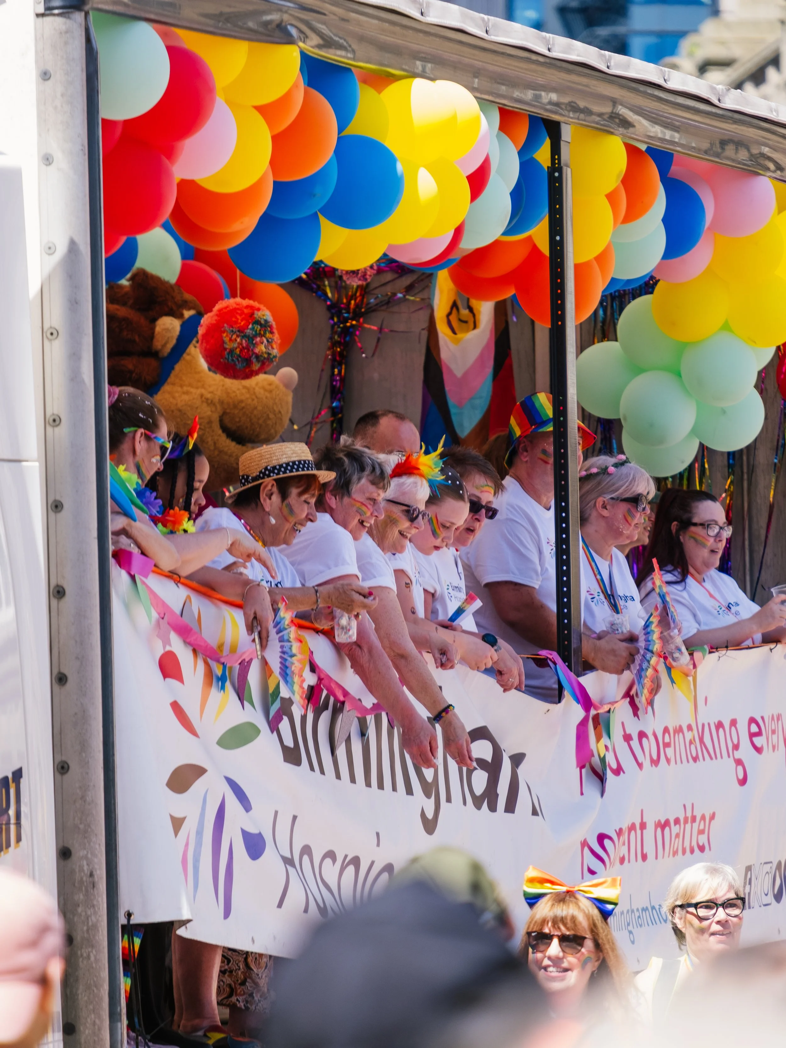 Participants in a pride parade standing behind a white banner with rainbow-colored decorations and text, decorated with rainbow flags, balloons in rainbow colors, and wearing rainbow-themed accessories.