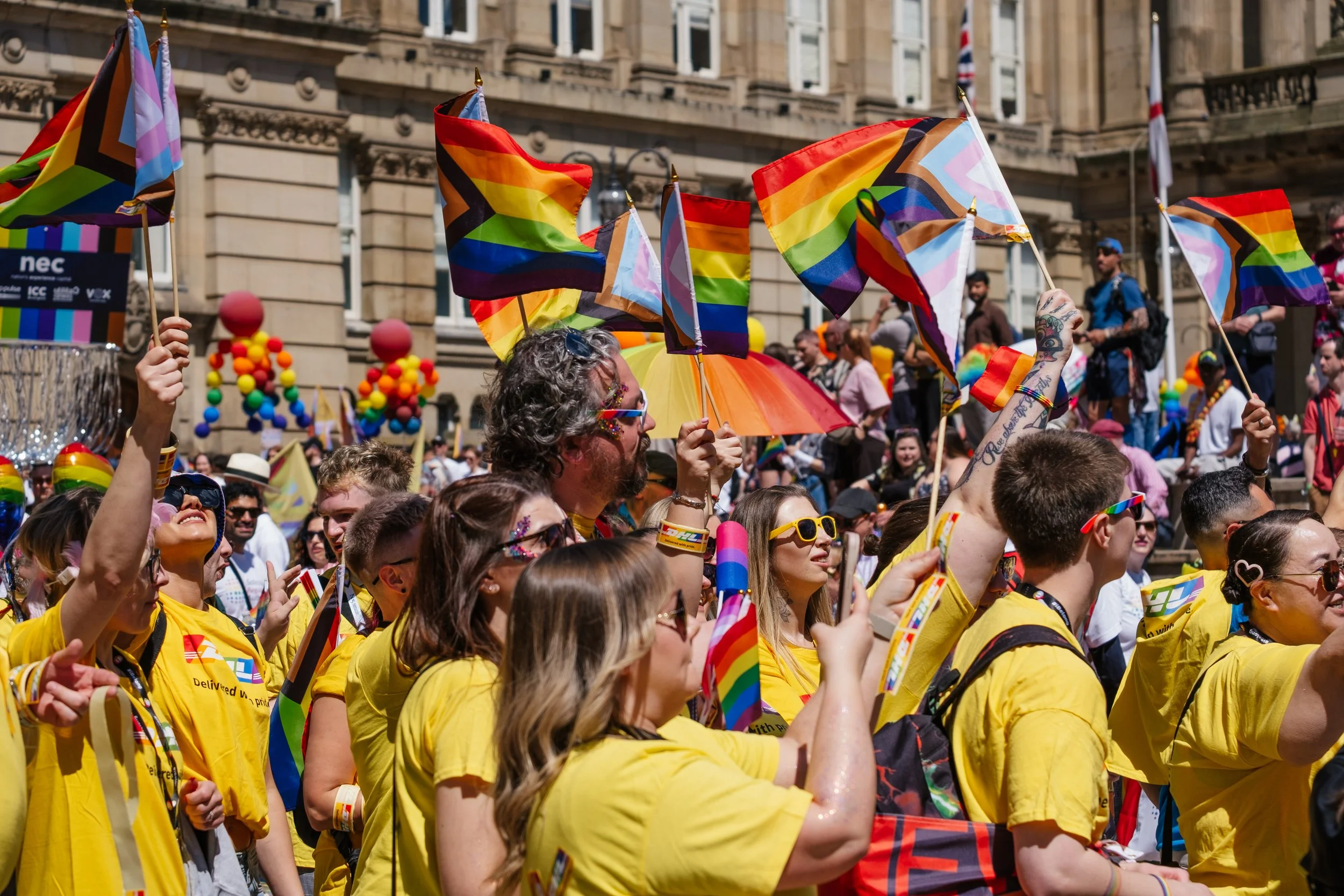 Crowd of people at Pride parade holding rainbow flags and wearing yellow shirts.