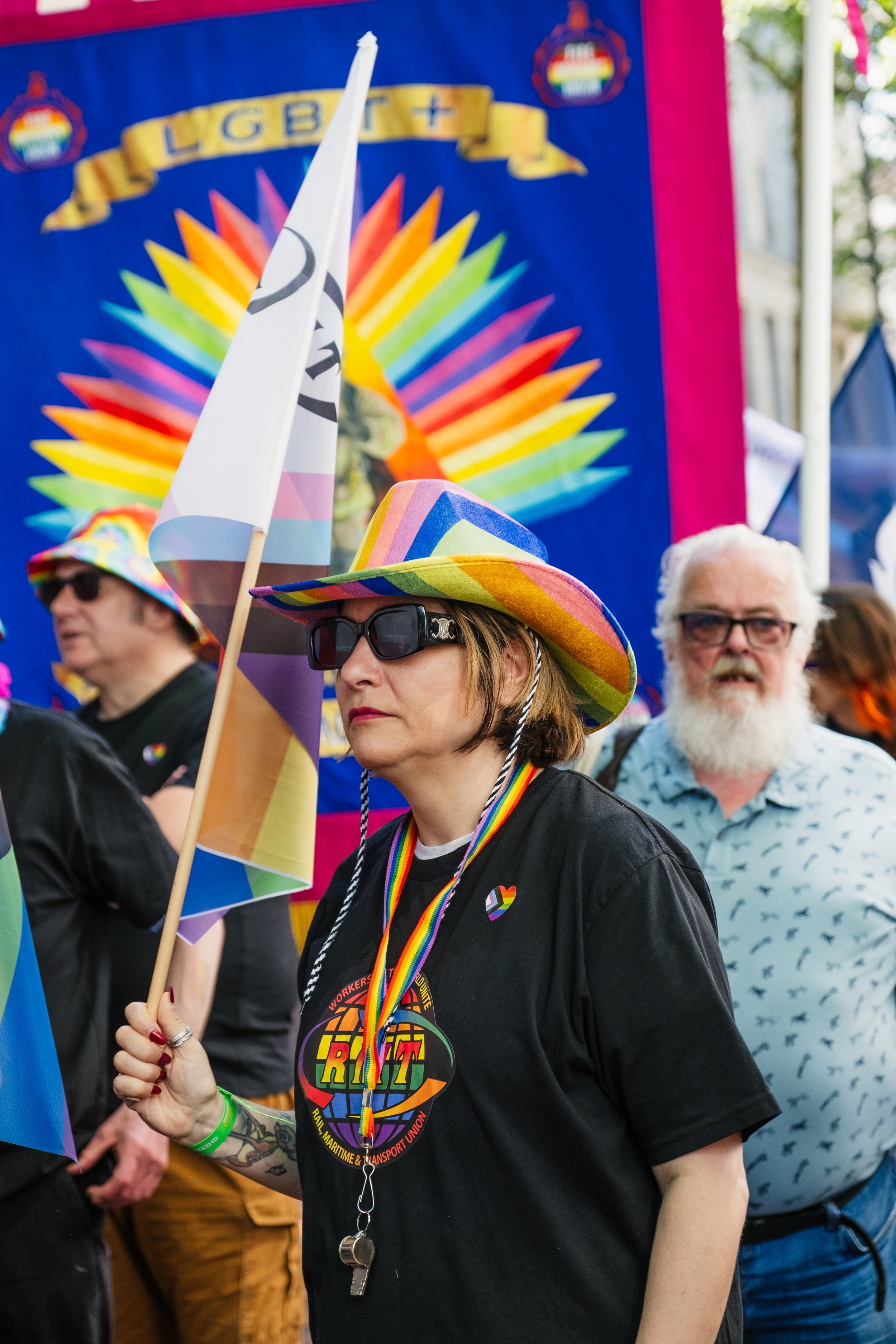 Women participating in a pride parade, wearing rainbow-themed accessories including a hat and bracelet, holding a rainbow flag, with a colorful pride banner in the background.