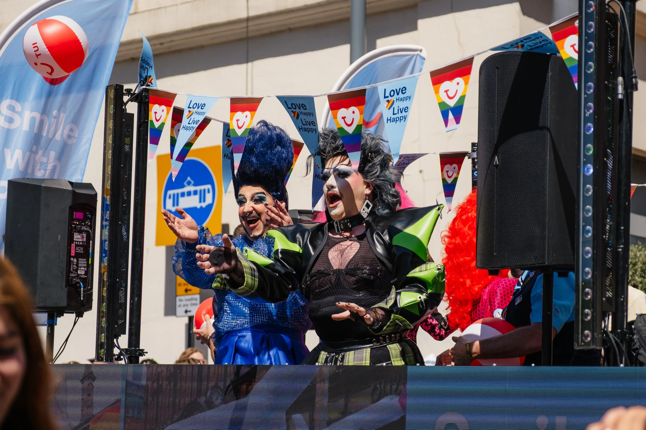 People in colorful costumes celebrating at an outdoor Pride parade, with banners and flags displaying messages of love, happiness, and rainbow pride.
