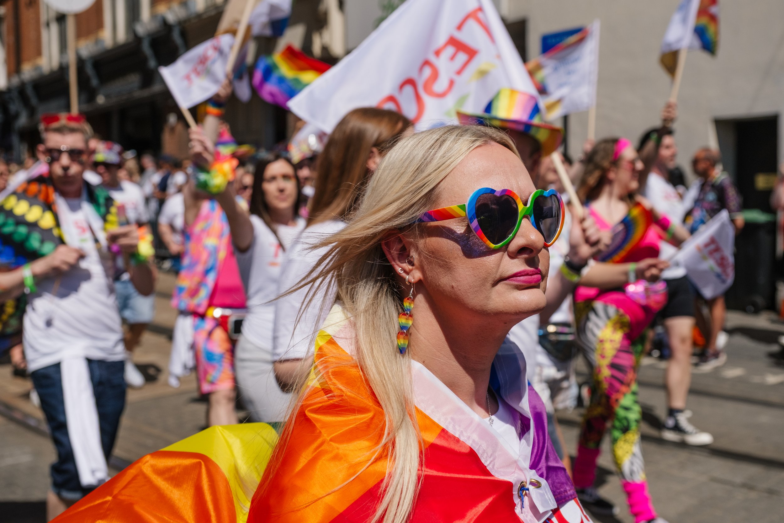 A woman with blonde hair wearing rainbow-colored heart-shaped sunglasses and rainbow earrings at a pride parade, with other parade participants in the background holding flags and wearing colorful outfits.