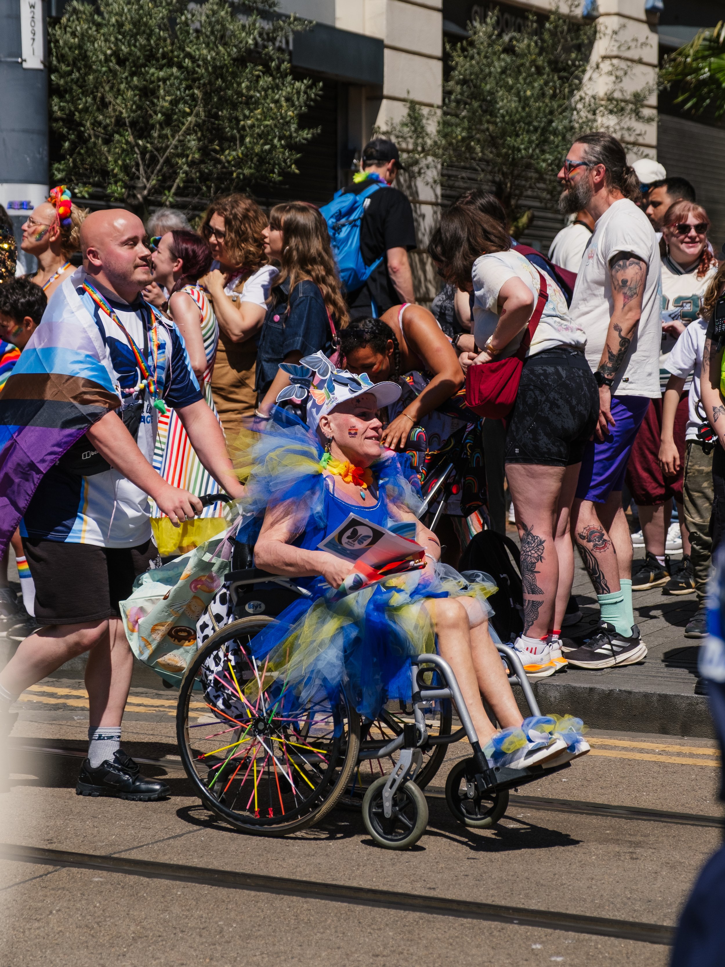 A group of people participating in a pride parade, including a person in a wheelchair wearing colorful rainbow-themed clothing and accessories, with rainbow decorations on the wheelchair. The crowd includes individuals wearing rainbow-themed attire, 