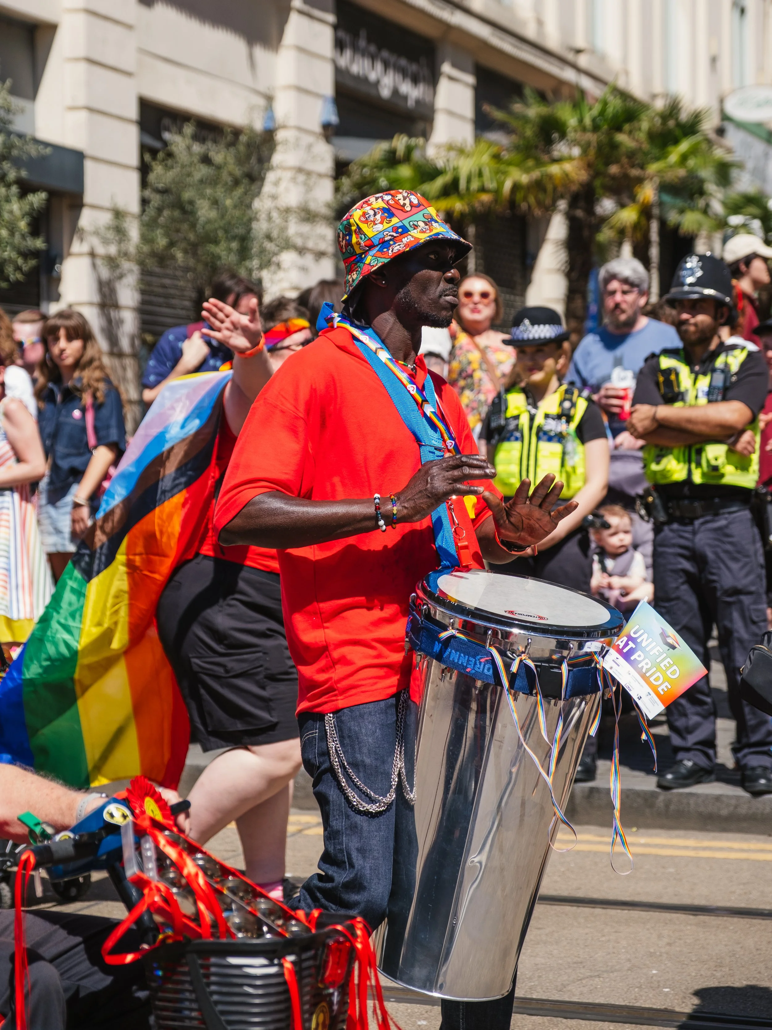 A man playing a drum at a pride parade, with a large crowd and police officers in the background.