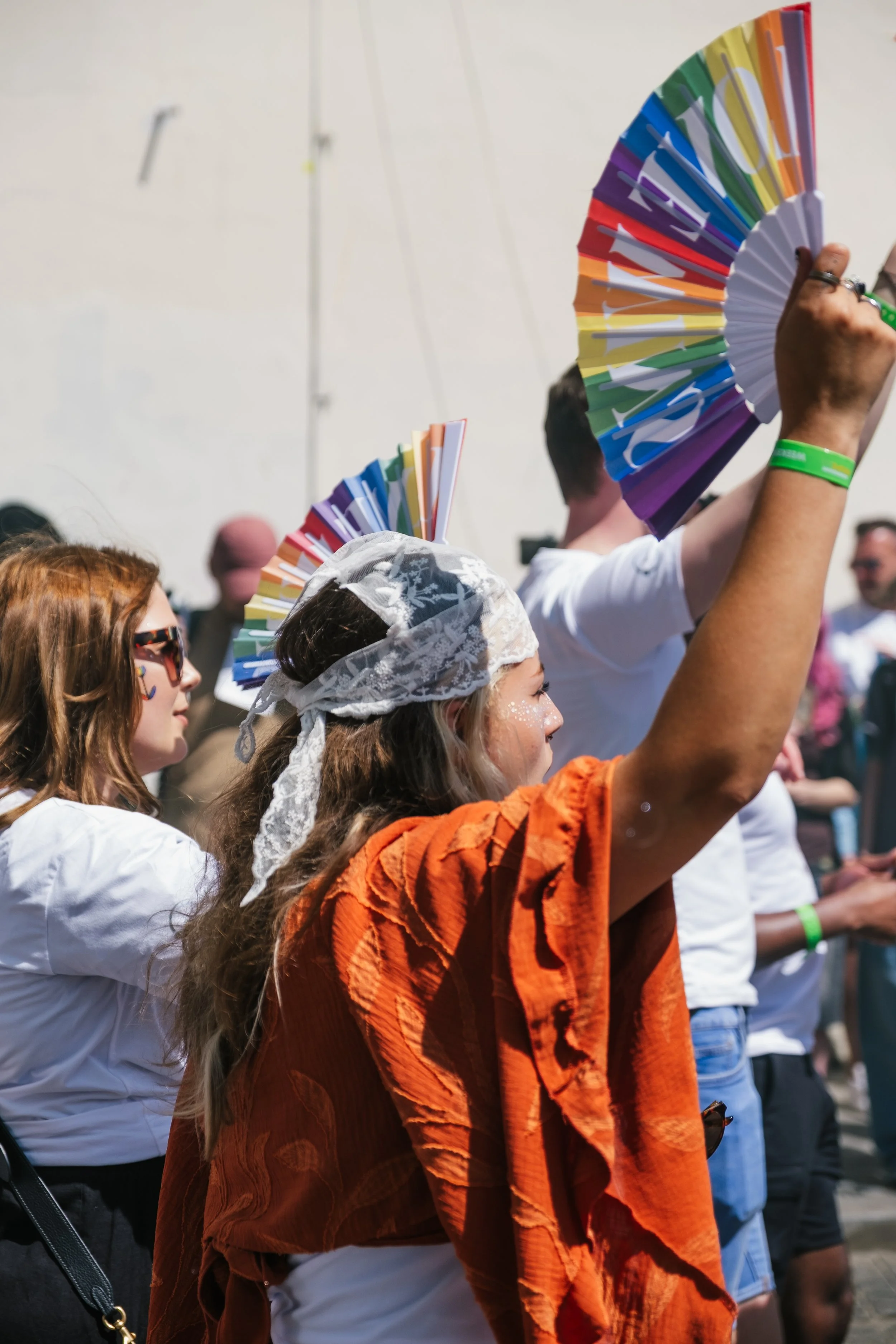 Several people at an outdoor event, holding rainbow-colored fans, with some wearing festival accessories like bandanas and wristbands.
