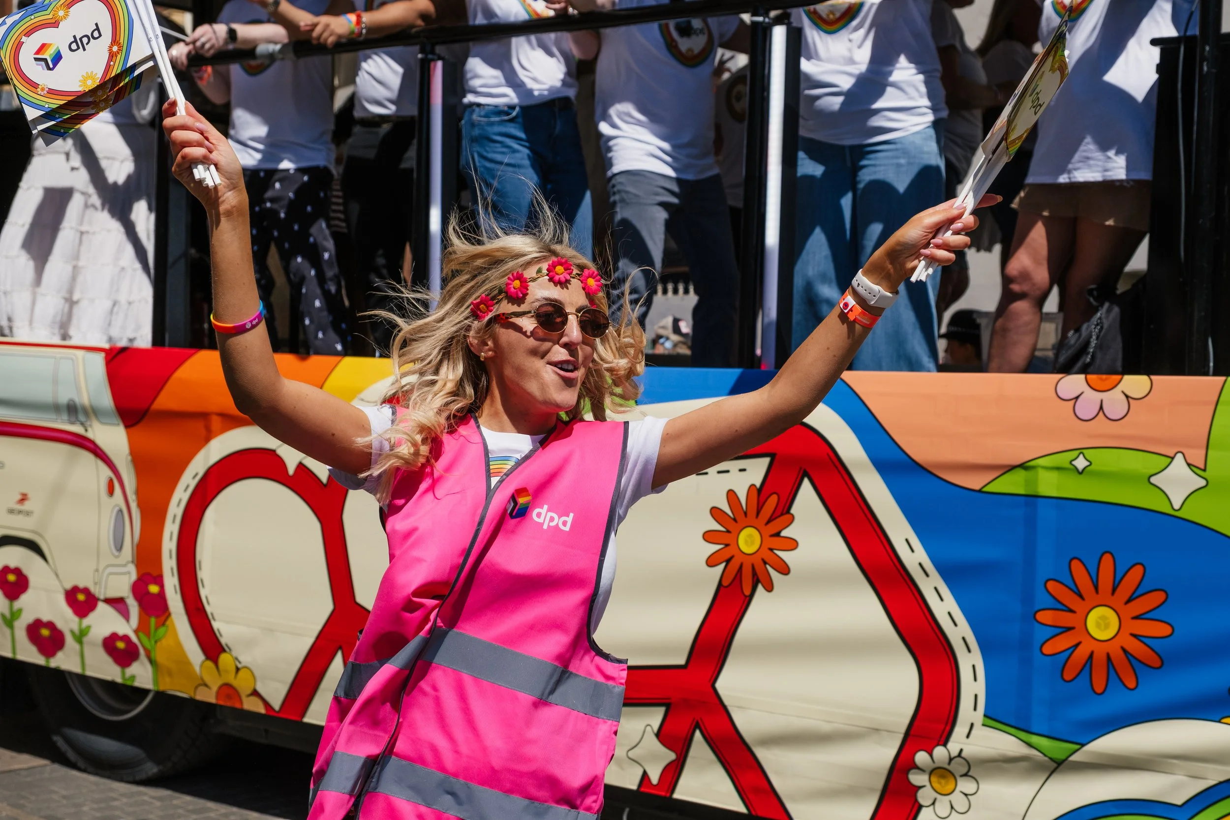 Woman with blonde hair, sunglasses, and a pink vest with a rainbow and flower design, waving and smiling during a parade or celebration, with a decorated float behind her.