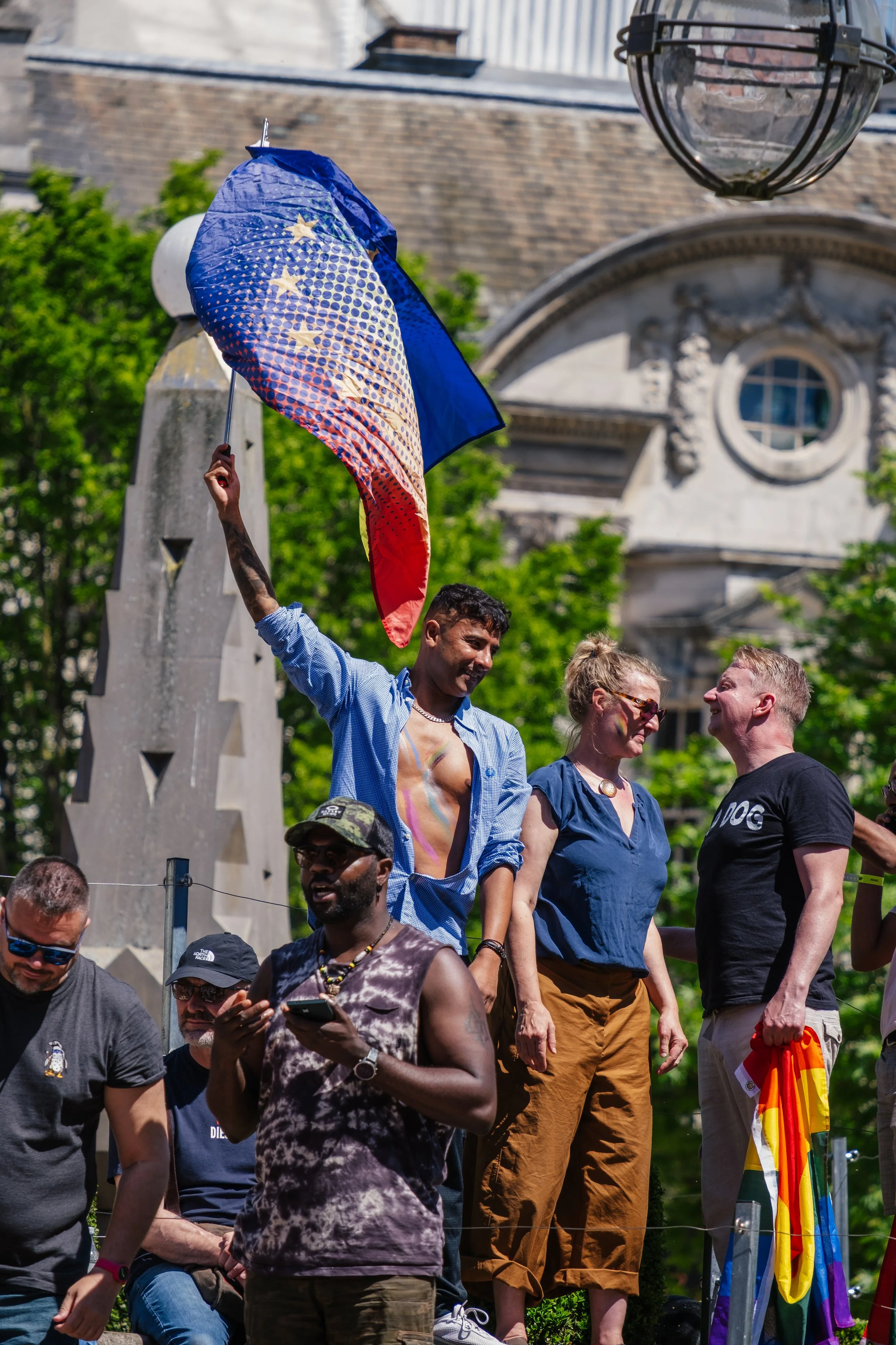 People gathered at an outdoor event, with a man holding a LGBTQ pride flag-shaped umbrella. The background features a historic building with architectural details, trees, and a rainbow Pride flag draped over a railing.