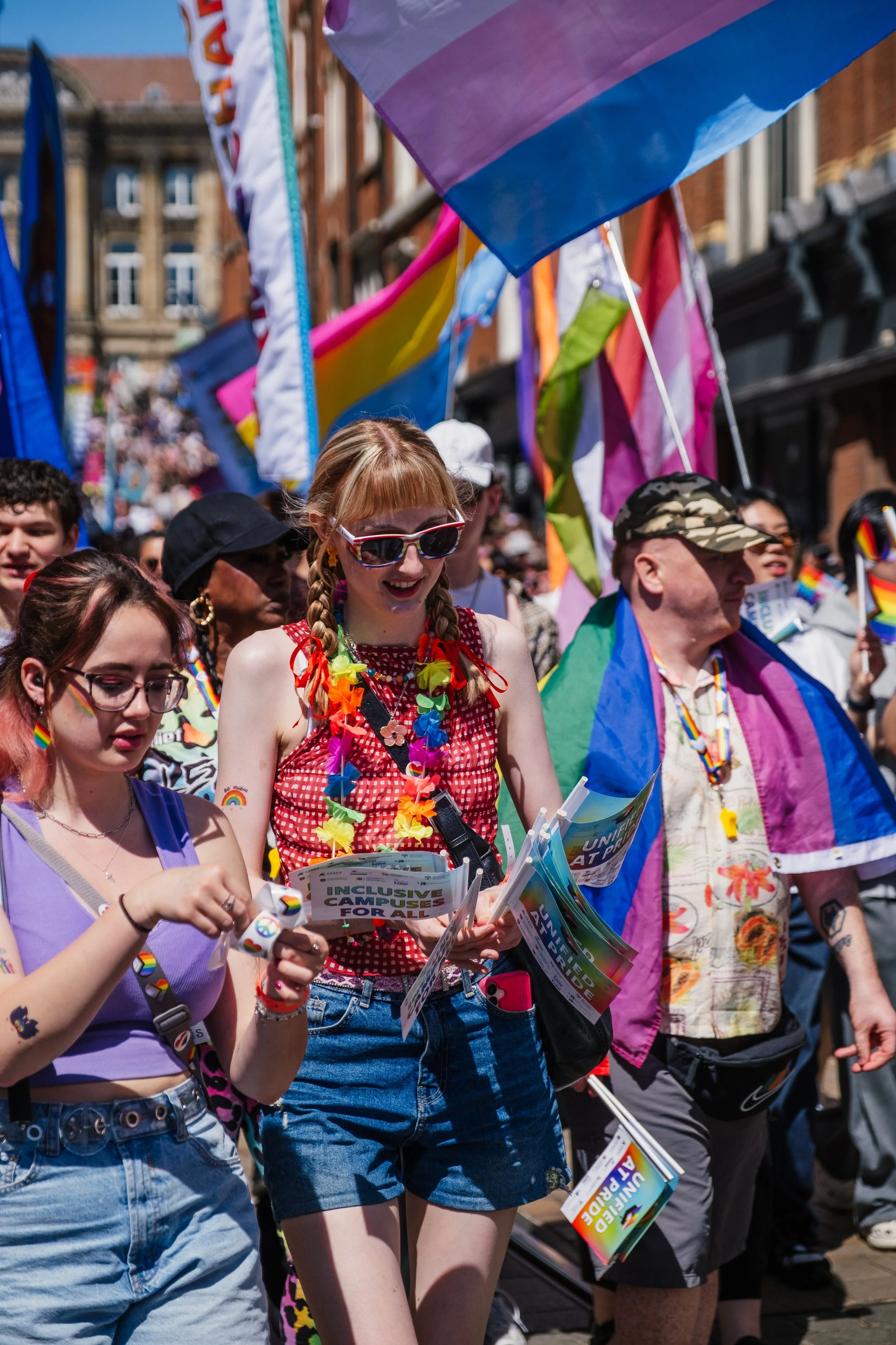 People participating in a pride parade, with rainbow flags and colorful clothing, walking on a city street under a clear blue sky.