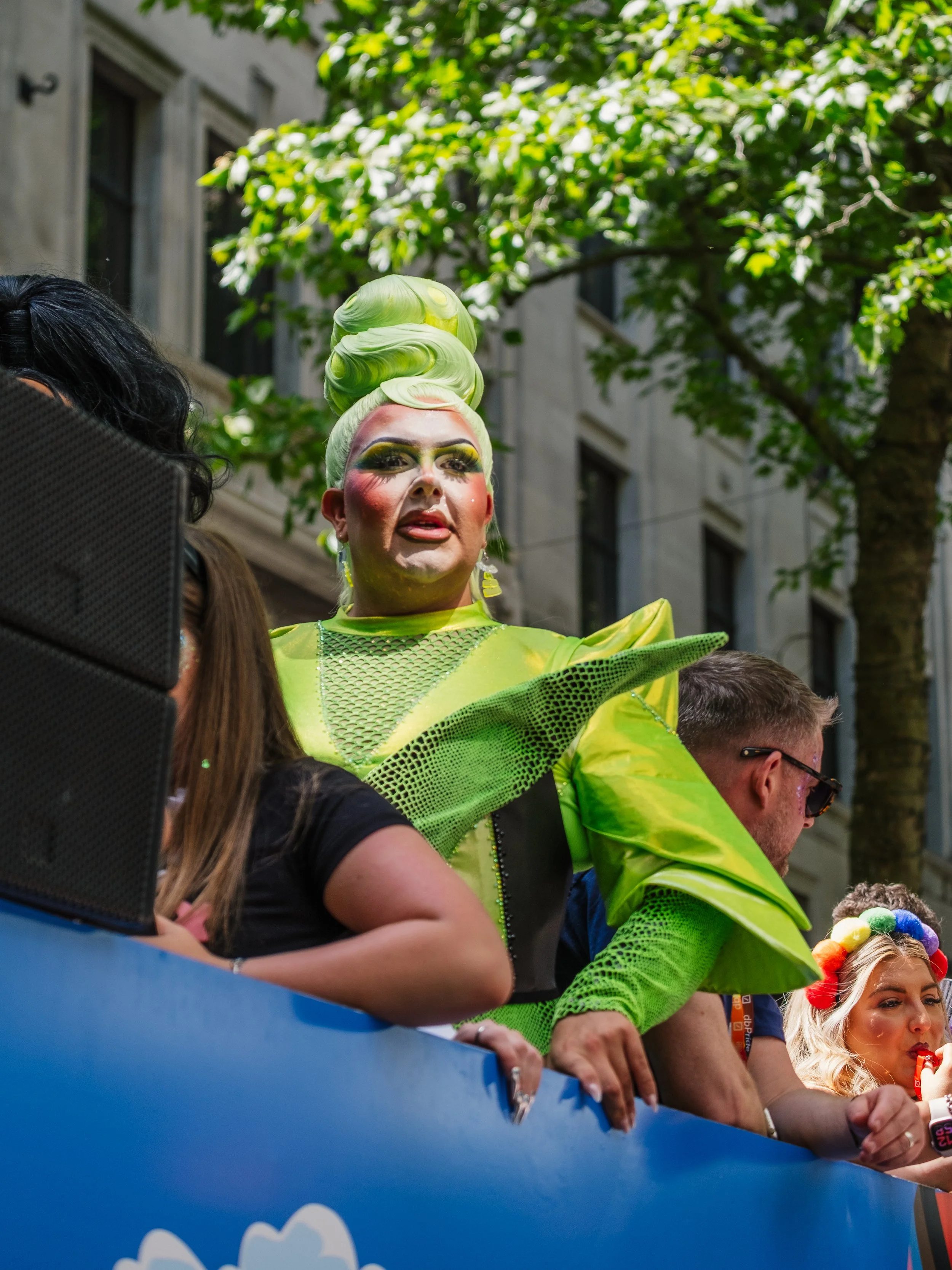 A person dressed in bright green, makeup, and elaborate costume, standing among others at a parade or festival, with a city building and green trees in the background.