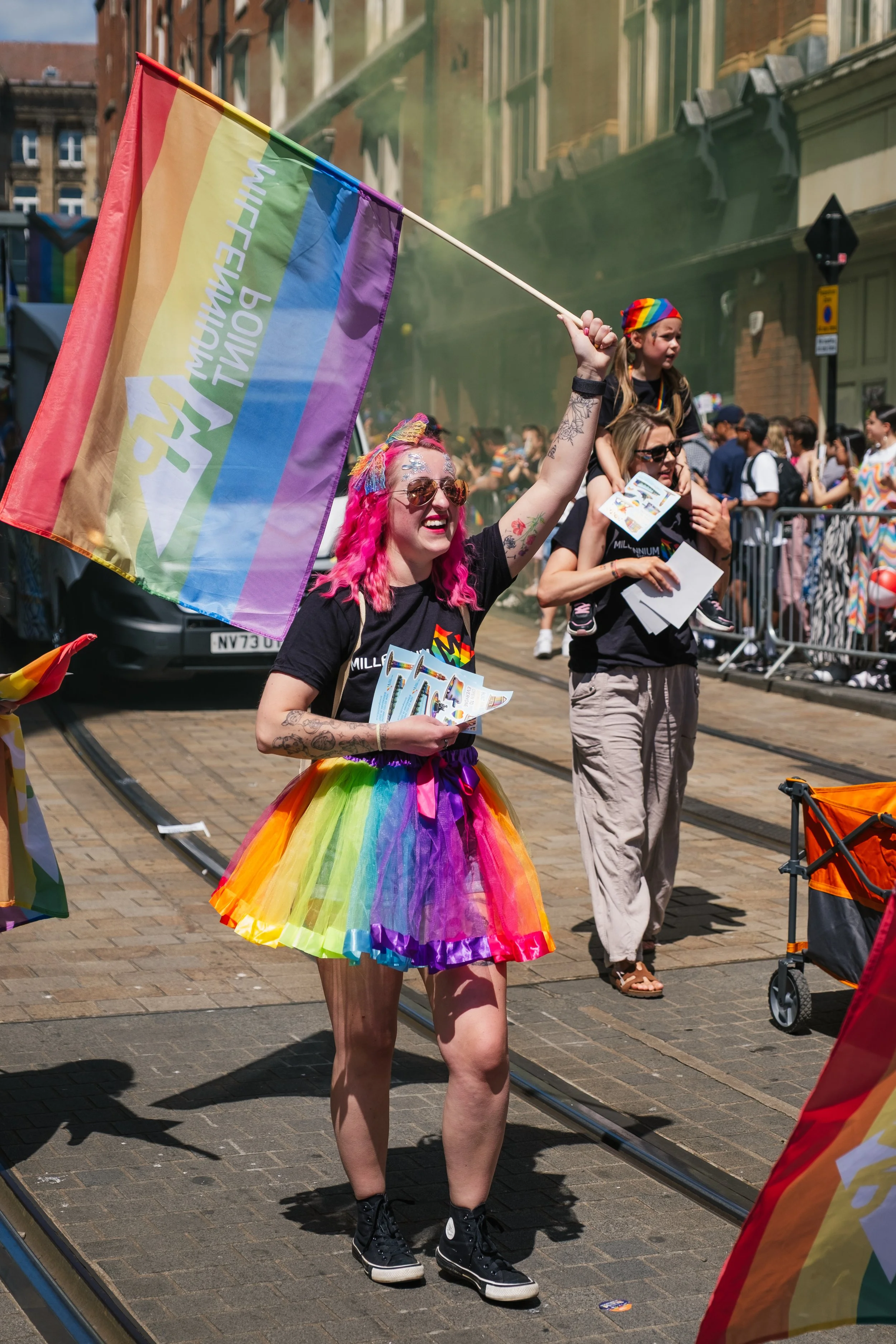 A person with pink hair and tattoos is smiling while walking in a pride parade, wearing a rainbow tutu, black t-shirt, sunglasses, and black sneakers. They hold a rainbow pride flag. A woman with a child on her shoulders, both wearing black t-shirts 