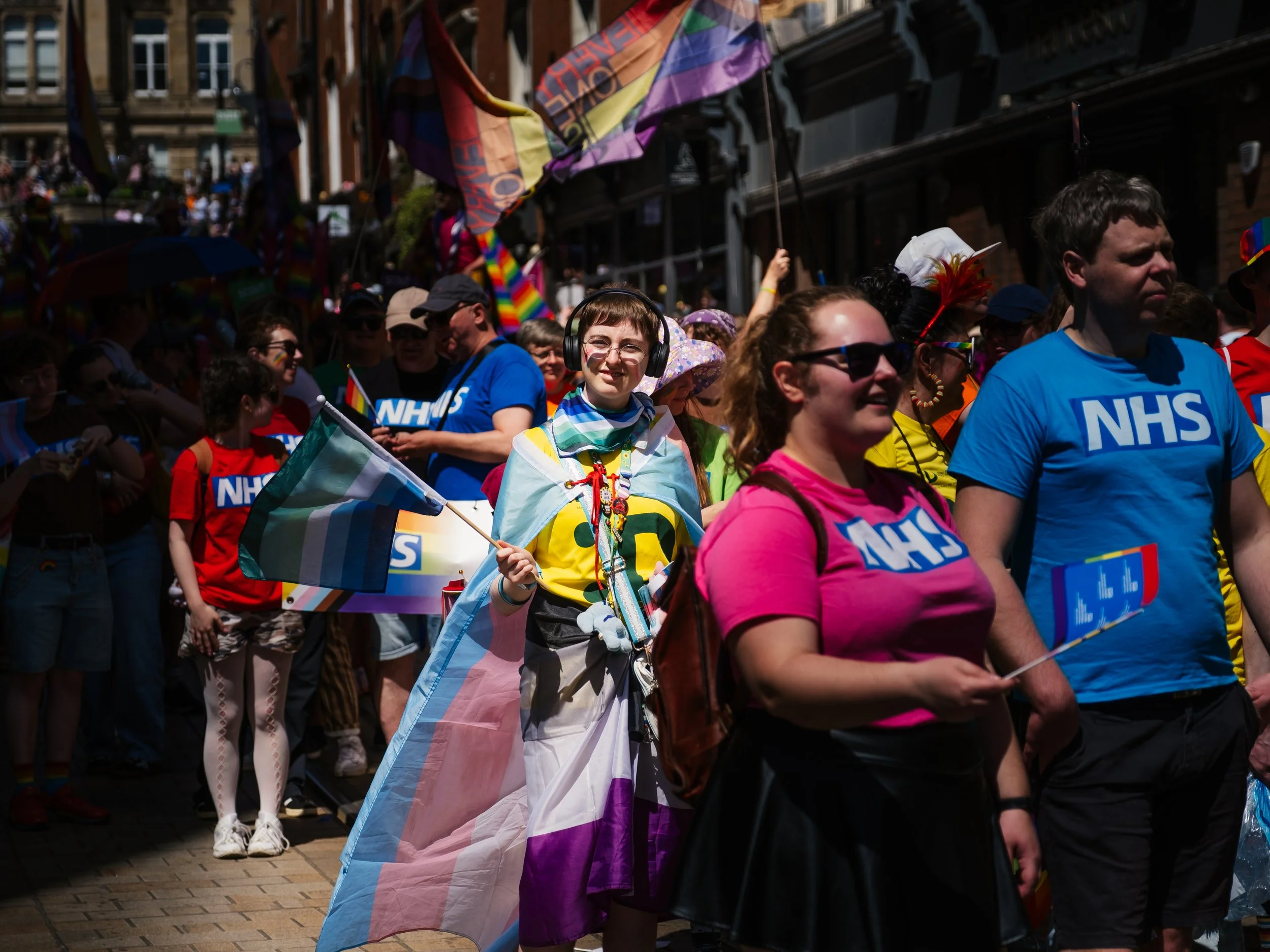 People participating in a pride parade, many wearing rainbow colors and holding flags, in an urban setting with buildings in the background.