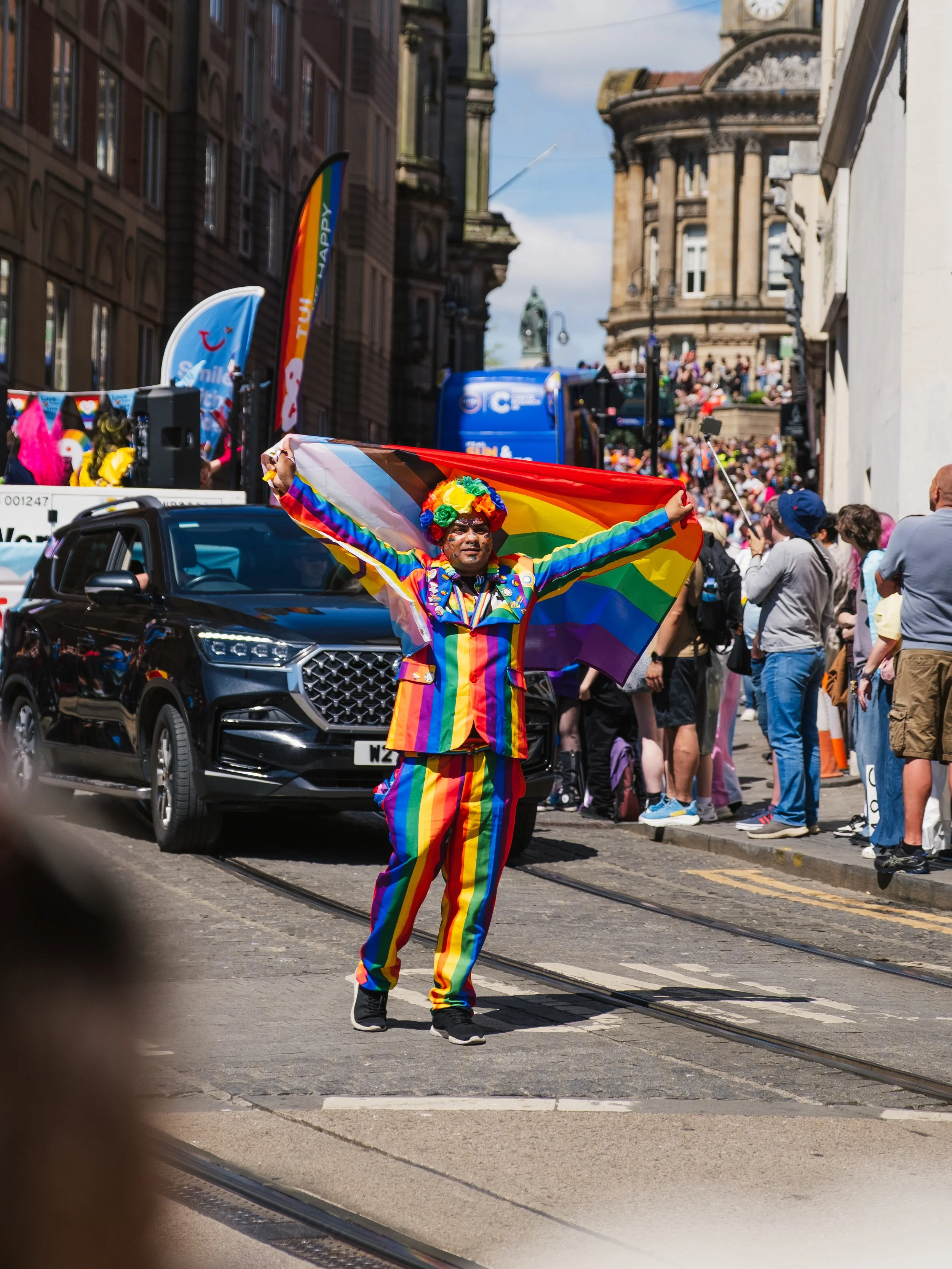 Person dressed in rainbow colors holding a rainbow pride flag at a parade or celebration, with a large crowd and buildings in the background.