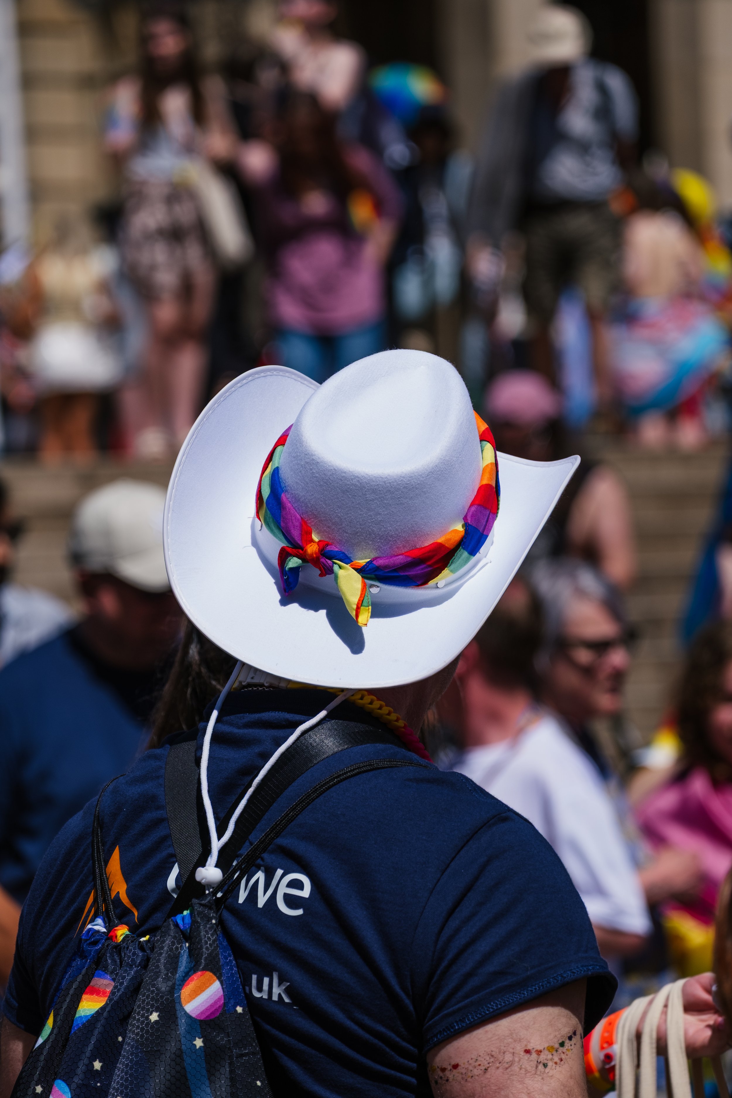 Person wearing a white cowboy hat with rainbow-colored band, dark shirt, and carrying a black bag with rainbow patterned straps at a crowded outdoor event.