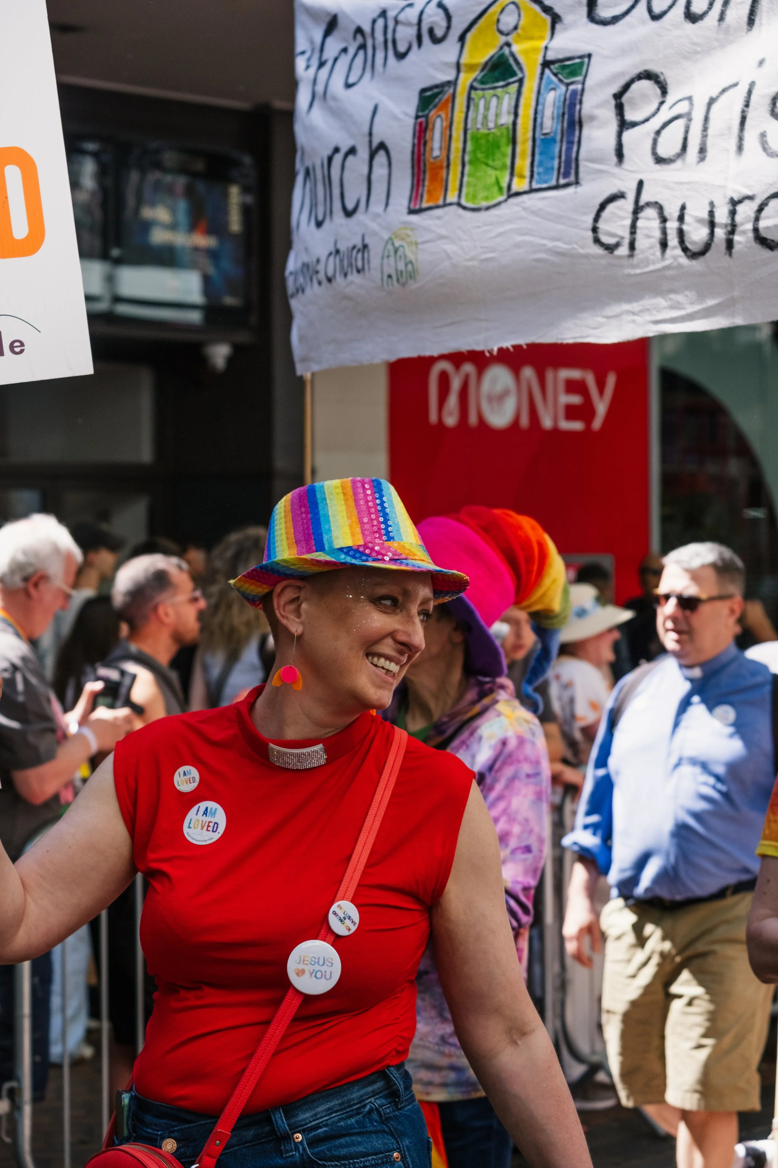 A diverse group of people participating in a parade or rally, with a woman smiling and wearing rainbow-colored accessories, buttons and a hat, surrounded by other individuals holding signs and wearing colorful clothing.