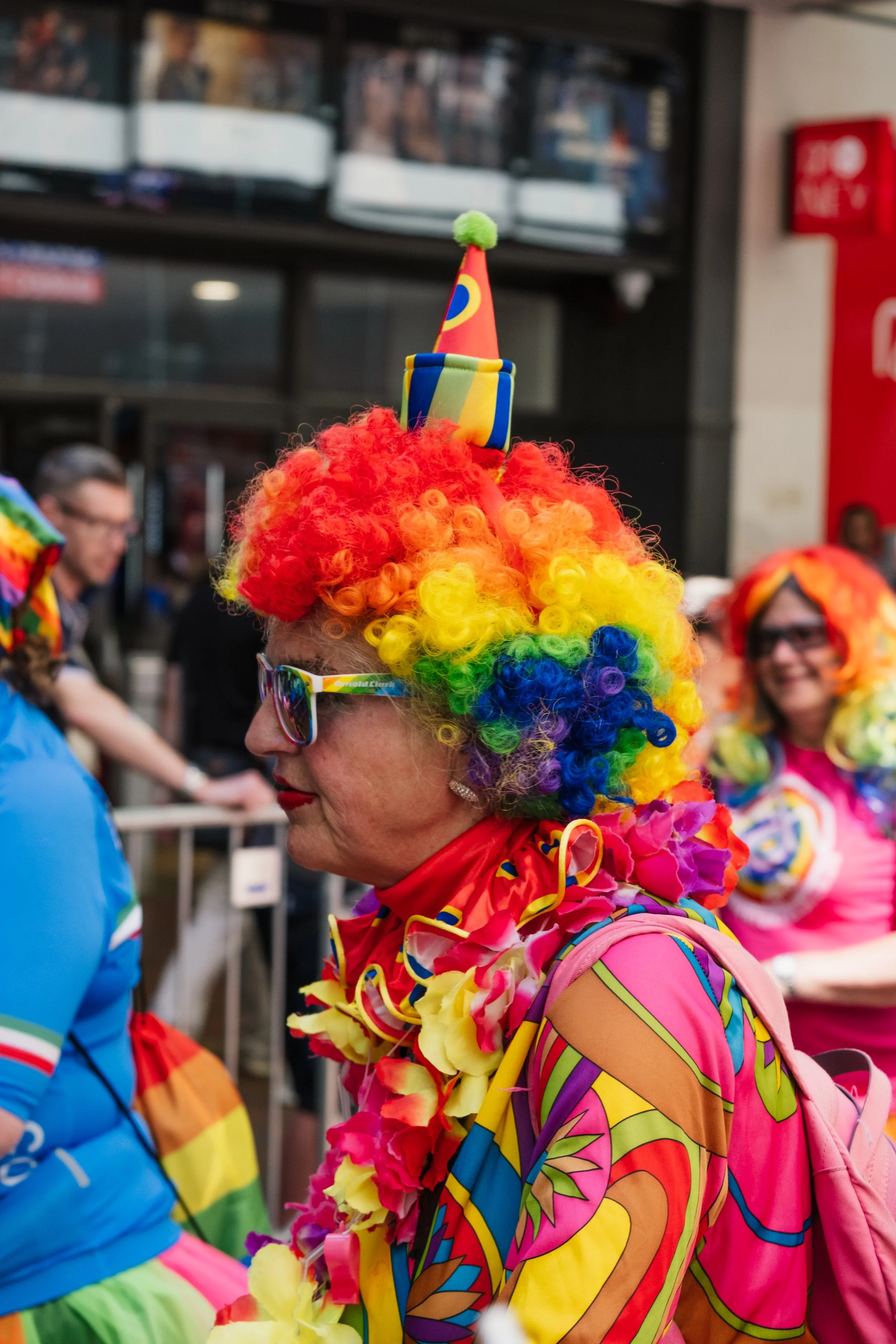 A woman wearing a colorful rainbow wig and a clown outfit, with a small clown hat on top, at a festive event, smiling and engaged in conversation. Background shows other attendees and an indoor setting.