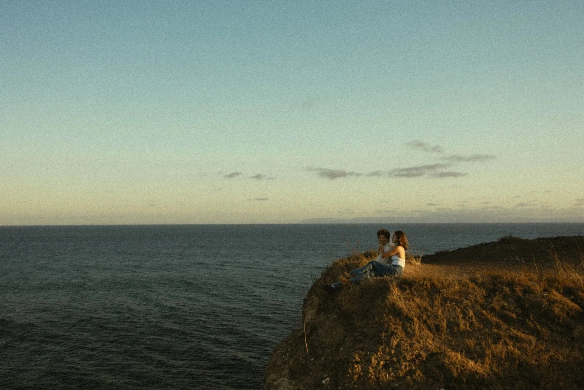 A couple sitting on a rocky cliff by the ocean during sunset, with the sky partially cloudy.