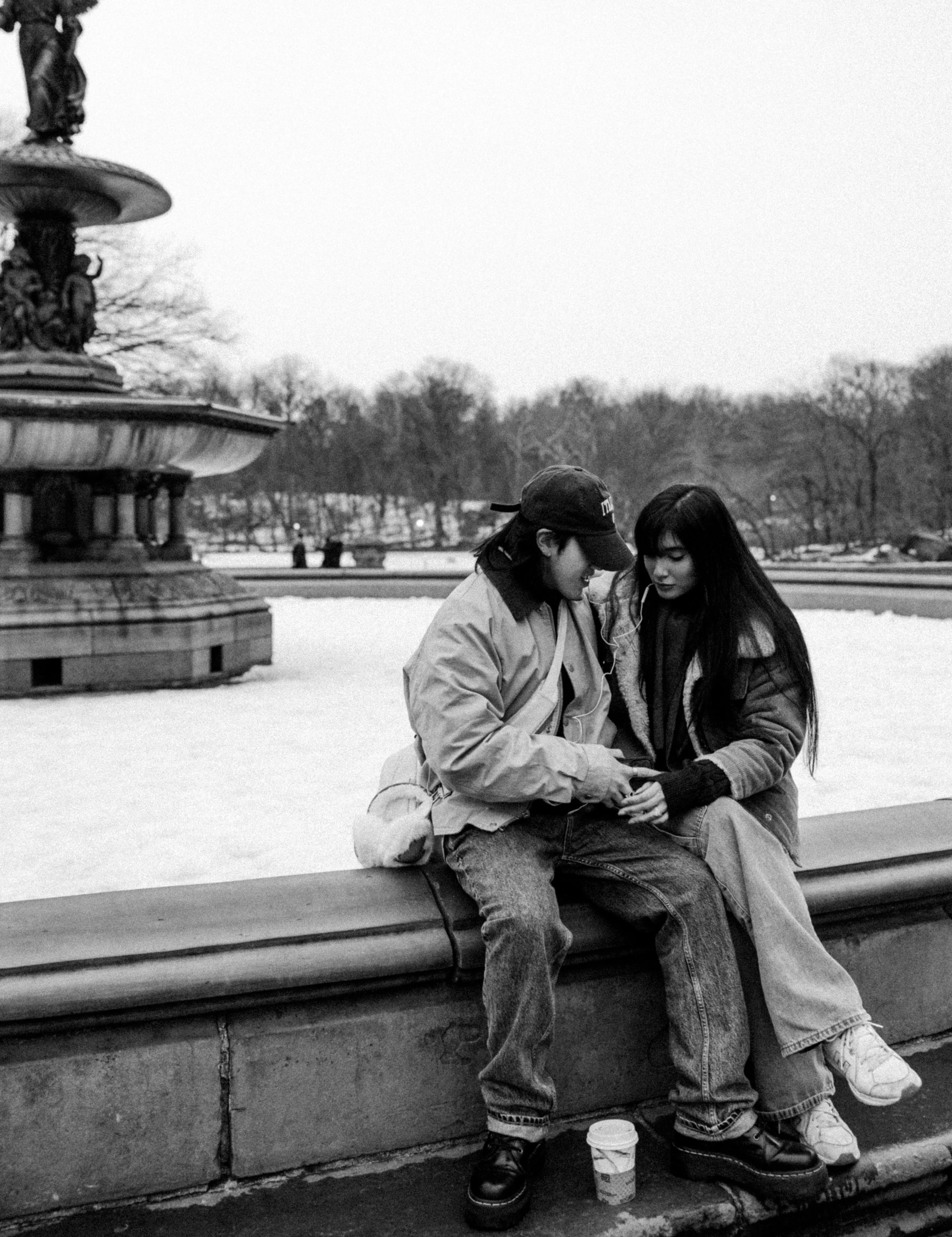 Two young women sitting on a park bench in black-and-white, engaging with a phone or device, with a large fountain in the background and trees lining the park.