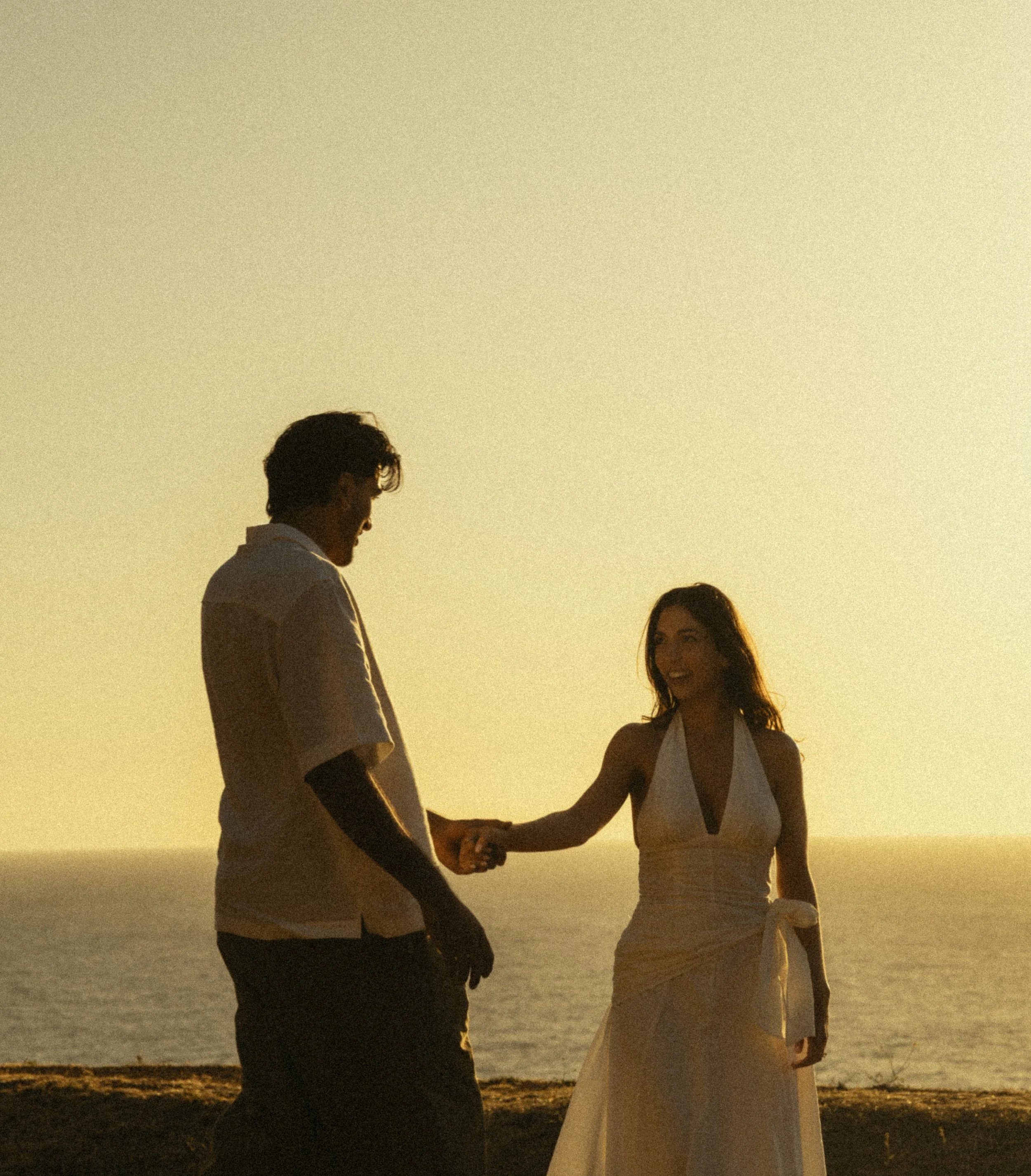 A man and woman holding hands and smiling at each other during sunset by the beach.