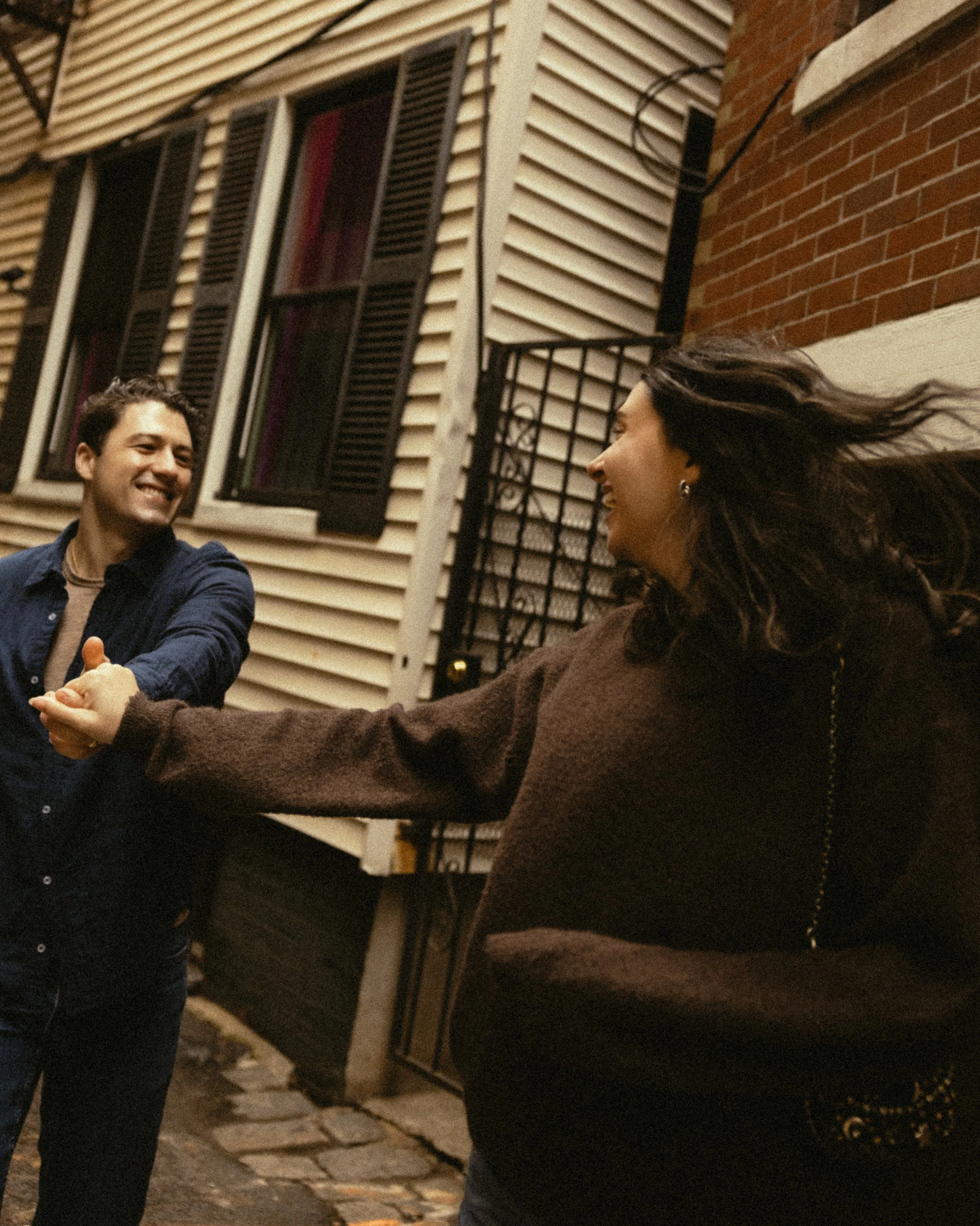 Two friends, a man and a woman, holding hands and smiling at each other outside at night with a house with open window shutters and brick wall in the background.
