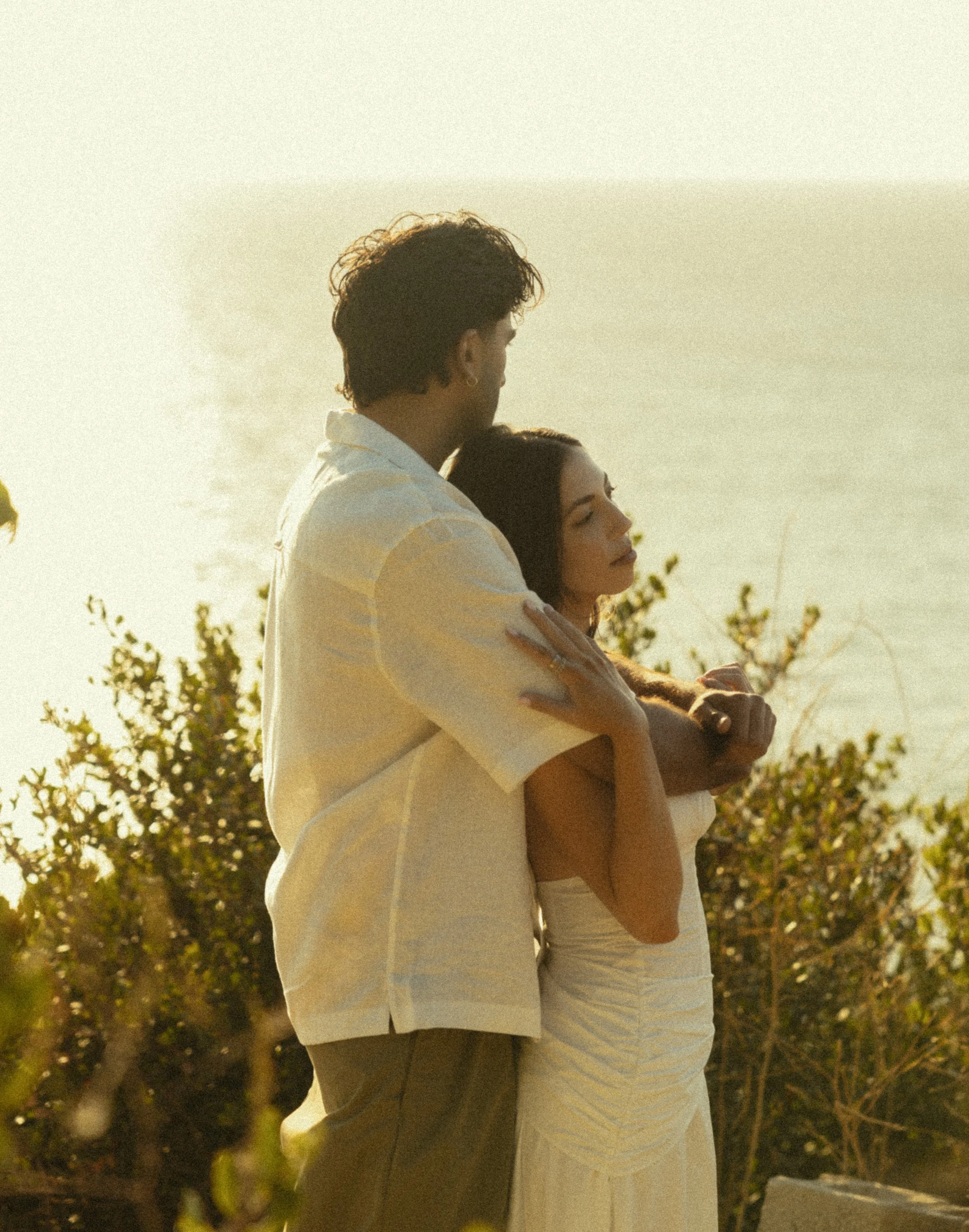 A man and woman hugging outdoors during sunset, with the man wearing a light-colored shirt and the woman in a white top, surrounded by trees.