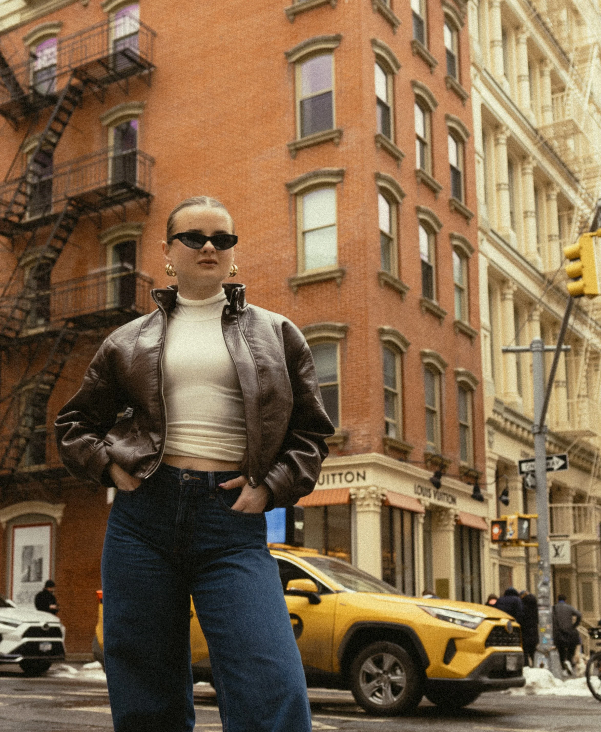 A woman with slicked-back hair wearing sunglasses, a beige turtleneck, and a brown leather jacket standing on a busy city street with yellow taxis, storefronts, and brick buildings in the background.