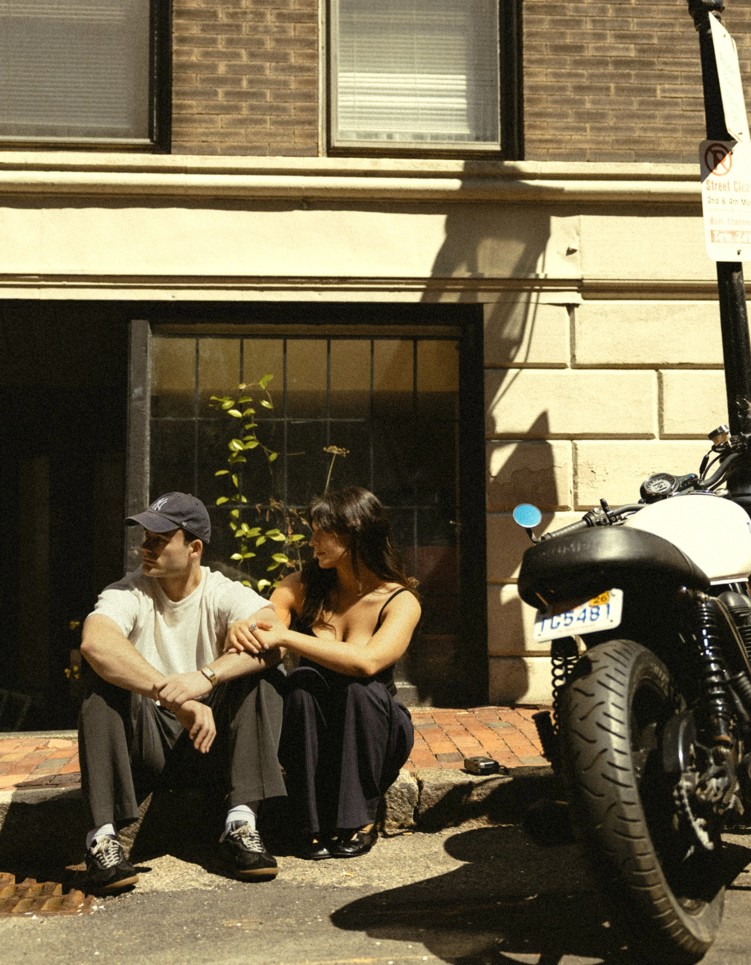 A young man and woman sit on a sidewalk at night, with the woman holding the man's hand. They are next to a motorcycle parked on the street, in front of a building with brick and stone walls and windows. The man is wearing a cap and sneakers, and the