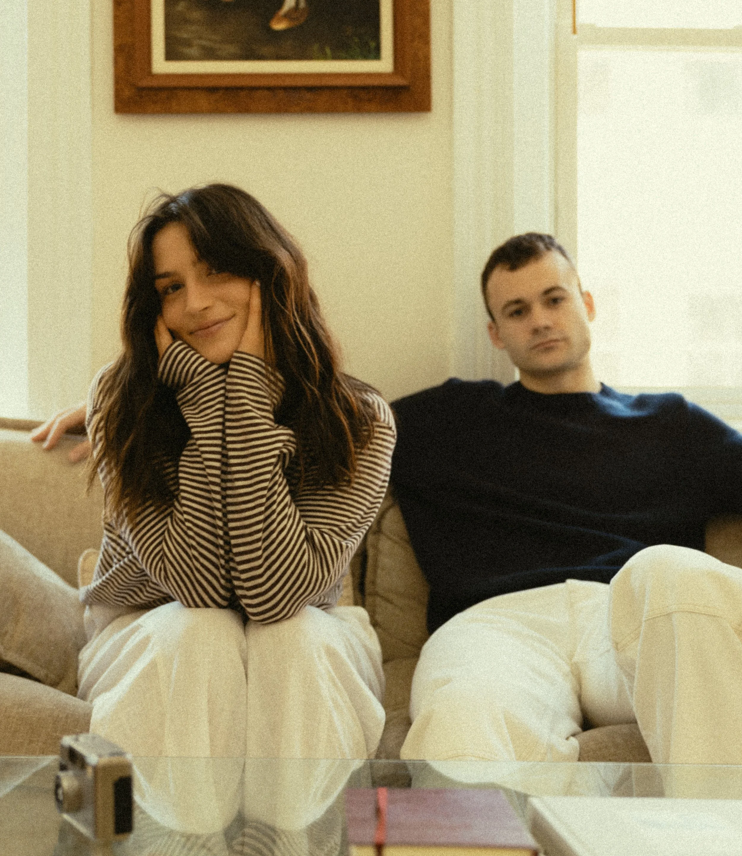 A woman with long dark hair smiling and resting her face on her hand, sitting on a beige couch, with a young man sitting beside her wearing a black shirt, in a room with a framed picture on the wall and a window.