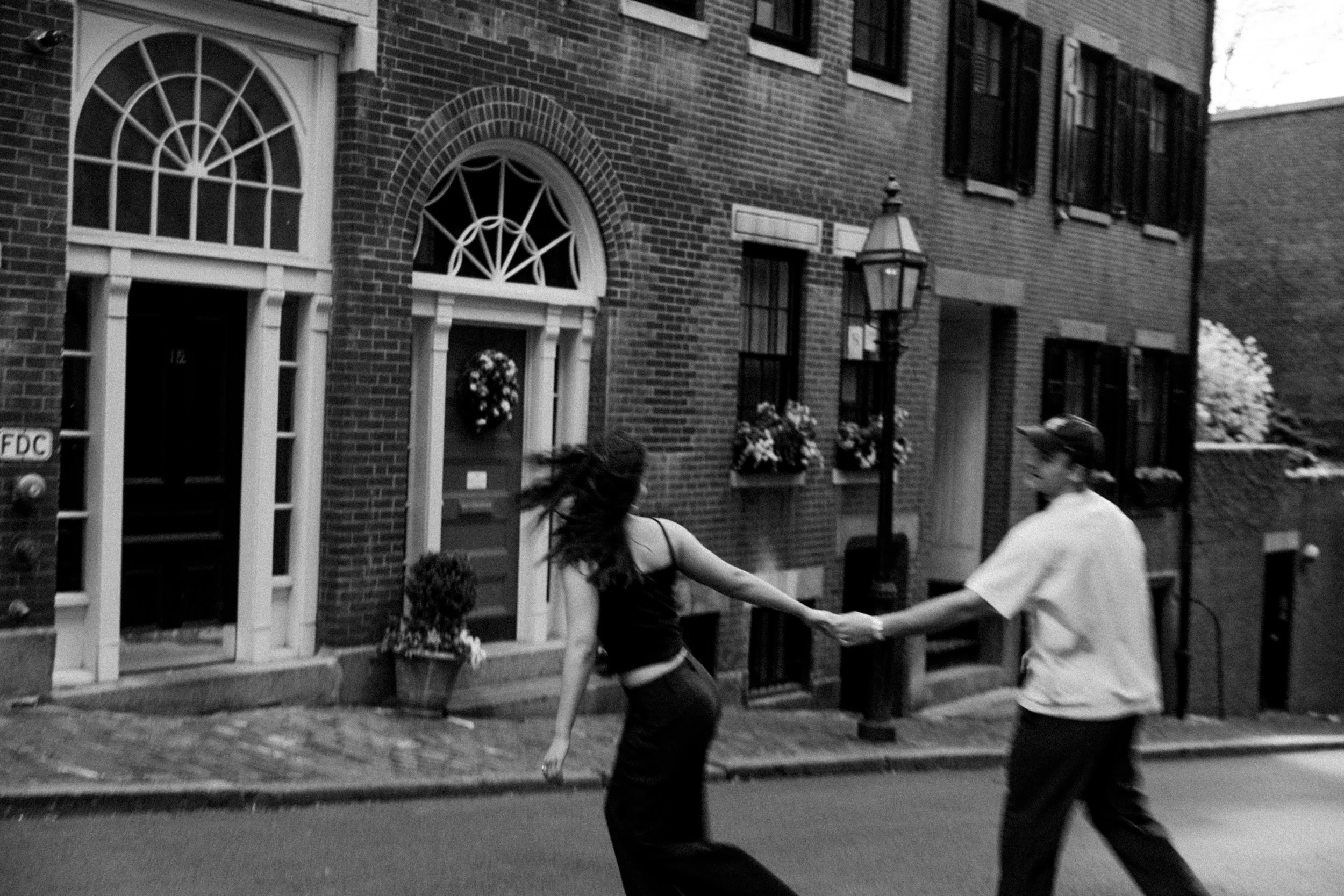 Two people holding hands and dancing or spinning on a city sidewalk in front of brick buildings with flowers and street lamps.