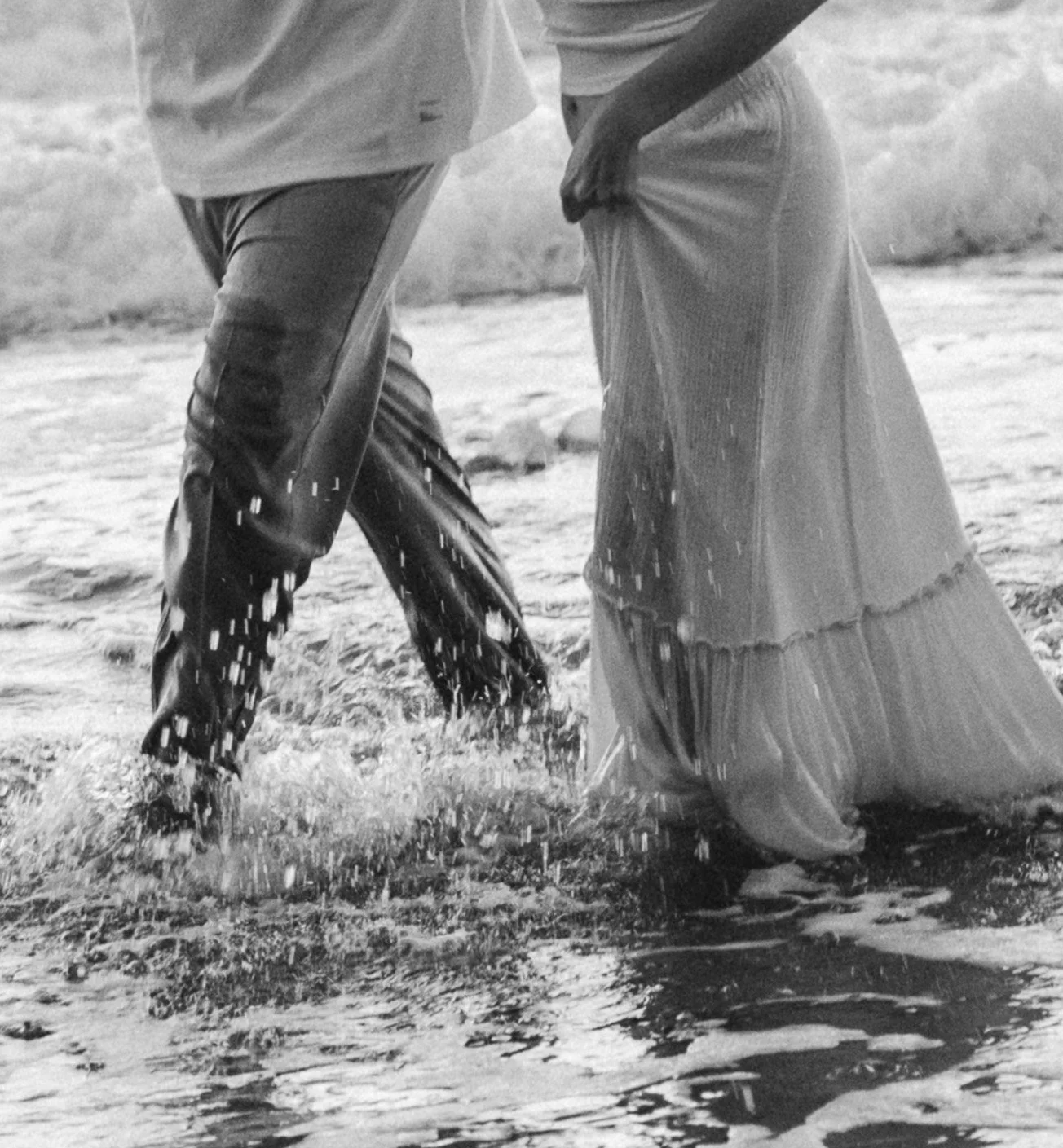 A black and white photo of two people standing in a shallow river with water splashing around them. One person is wearing dark pants and a light shirt, and the other is wearing a long, flowing skirt.
