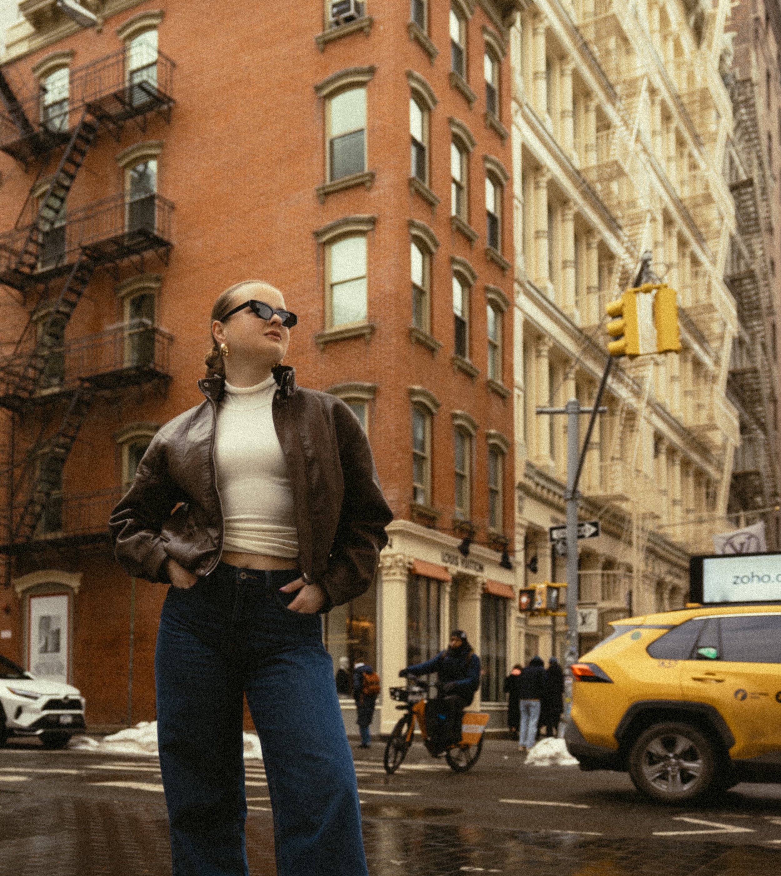 A woman wearing sunglasses, a brown leather jacket, and jeans standing on a city street with brick and beige buildings, a yellow taxi, a person on a bicycle, and other pedestrians.