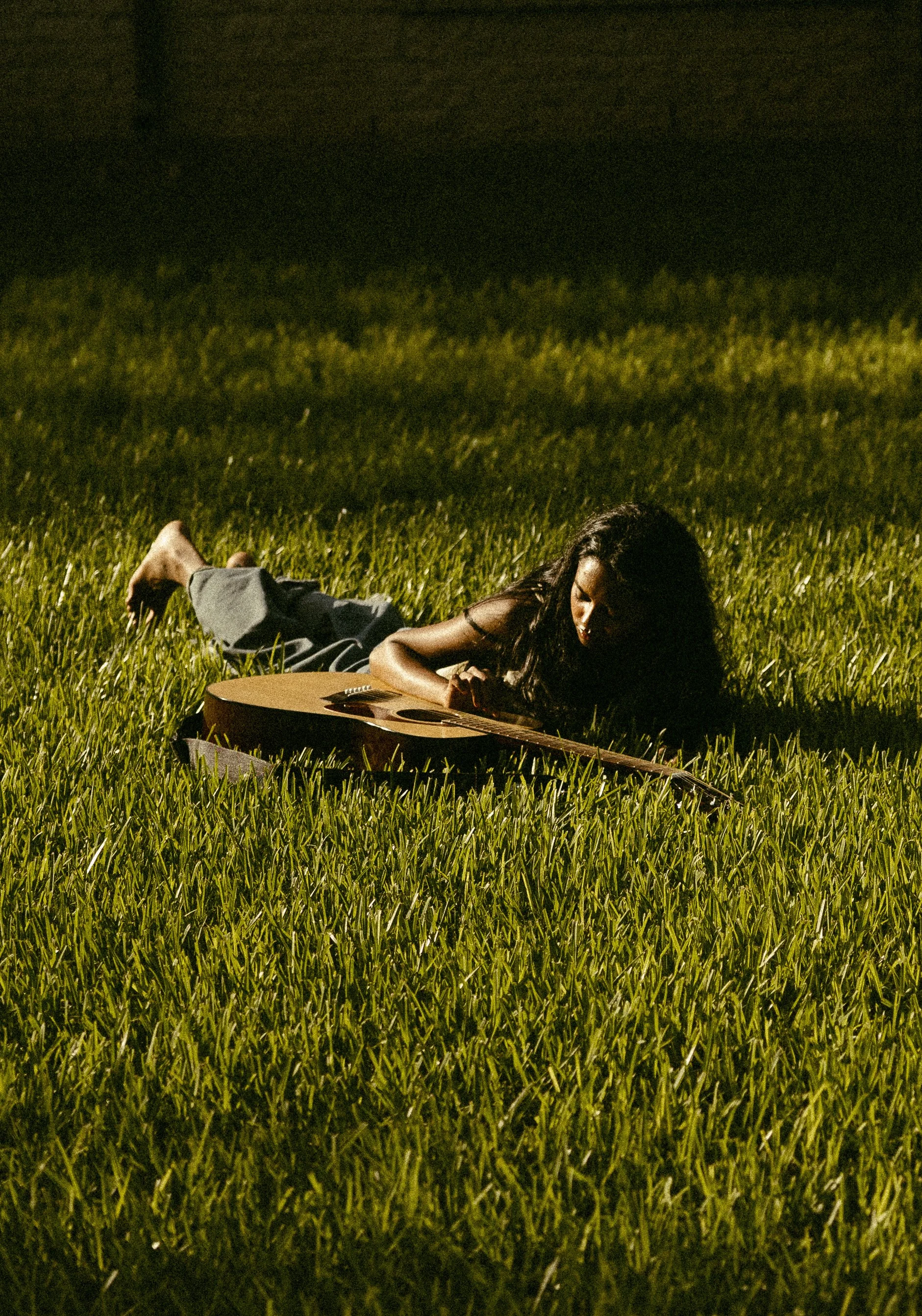A woman lying on her stomach on a grassy field at night, with a guitar resting in front of her.