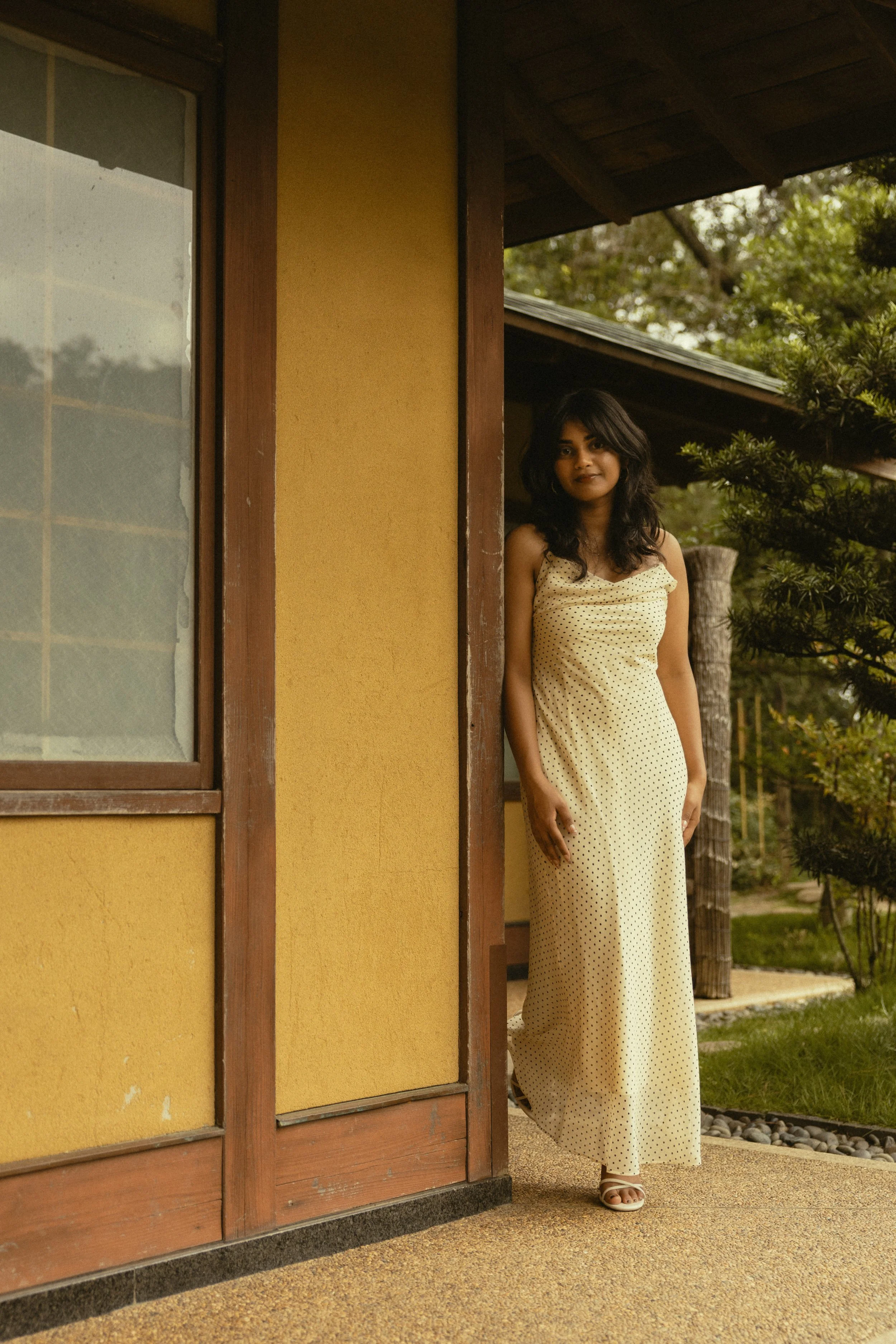 A woman with long dark hair wearing a cream-colored polka dot dress stands outside near a yellow wall and wooden window frame, with trees and greenery in the background.