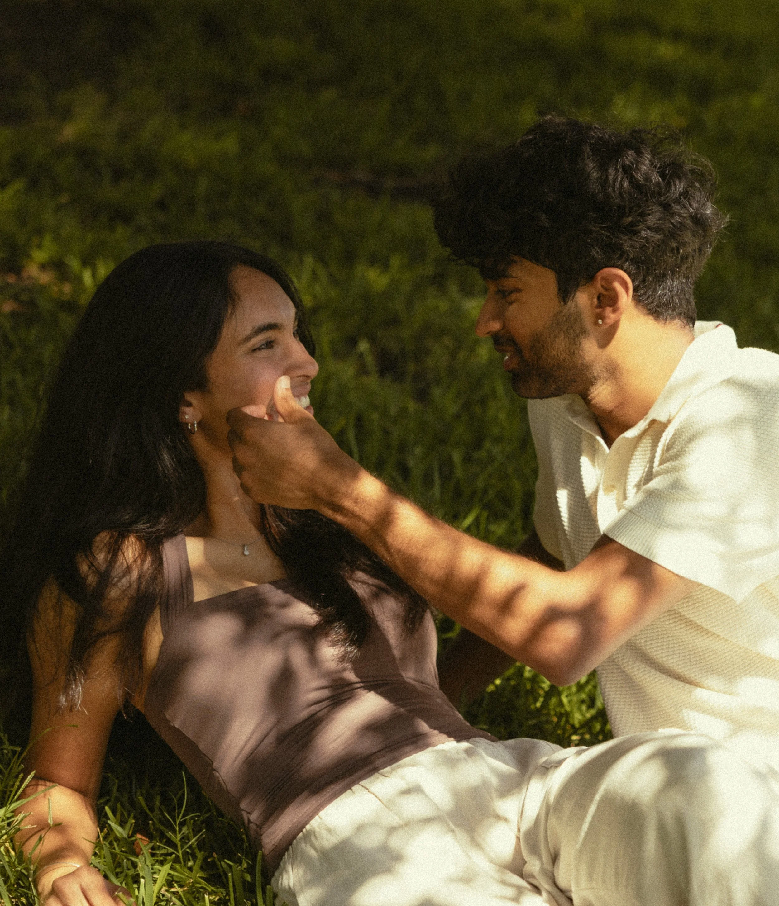 A young man and woman lie on grass facing each other, smiling and looking into each other's eyes.