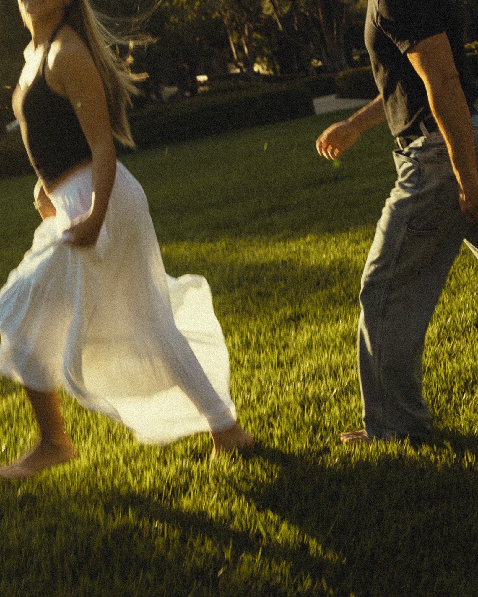 A woman wearing a black top and white skirt holding hands with a person wearing a black t-shirt and gray pants, outdoors at sunset on grass.