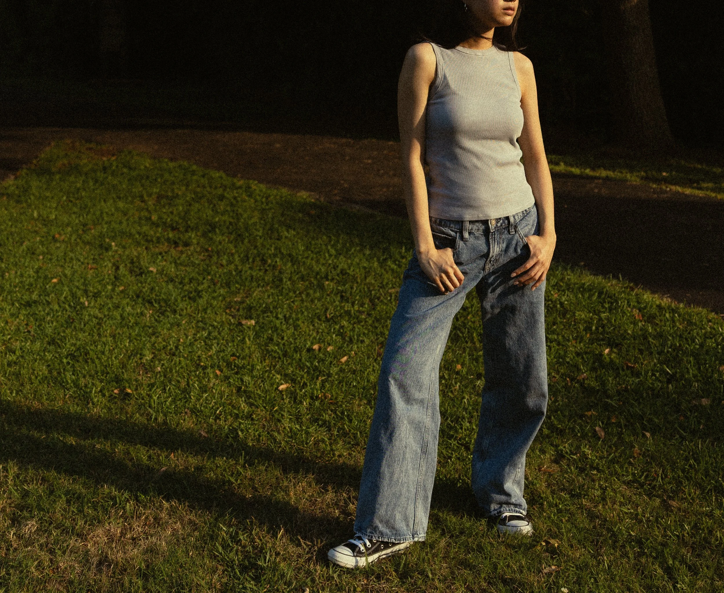 A person standing on a grassy area at night, wearing a sleeveless gray top, jeans, and sneakers, with only their lower face and torso visible.