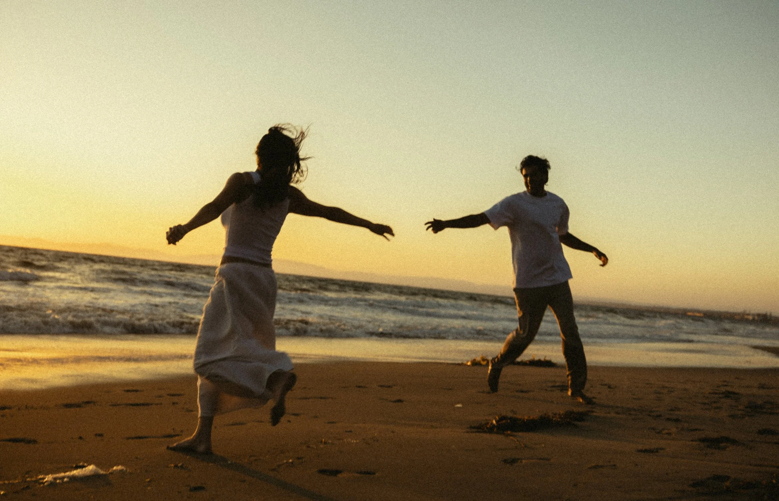 Two people holding hands and playing on the beach during sunset, with waves in the background and the sky illuminated by the setting sun.