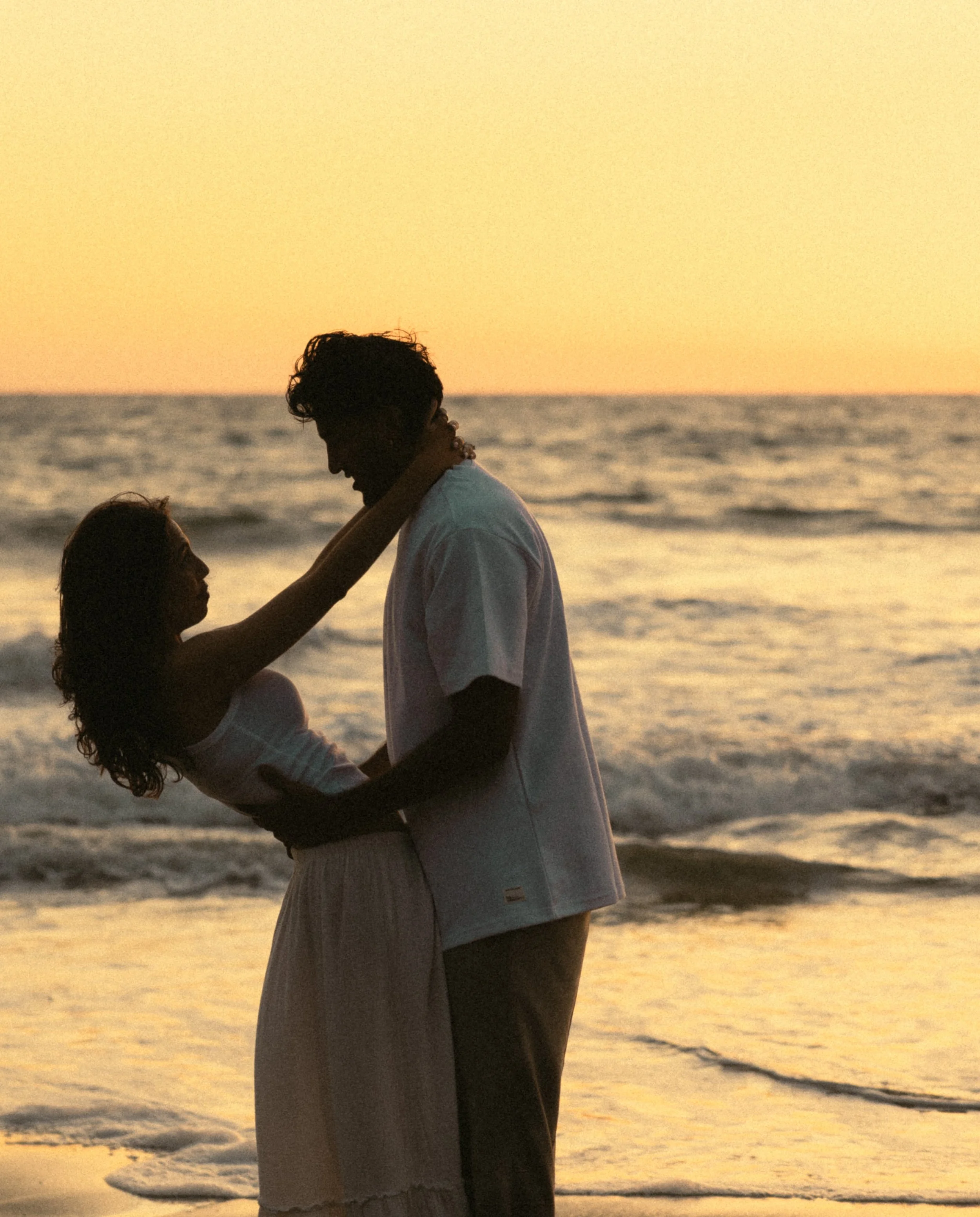 Silhouettes of two people, a man and a woman, on a beach at sunset, with the ocean waves in the background.