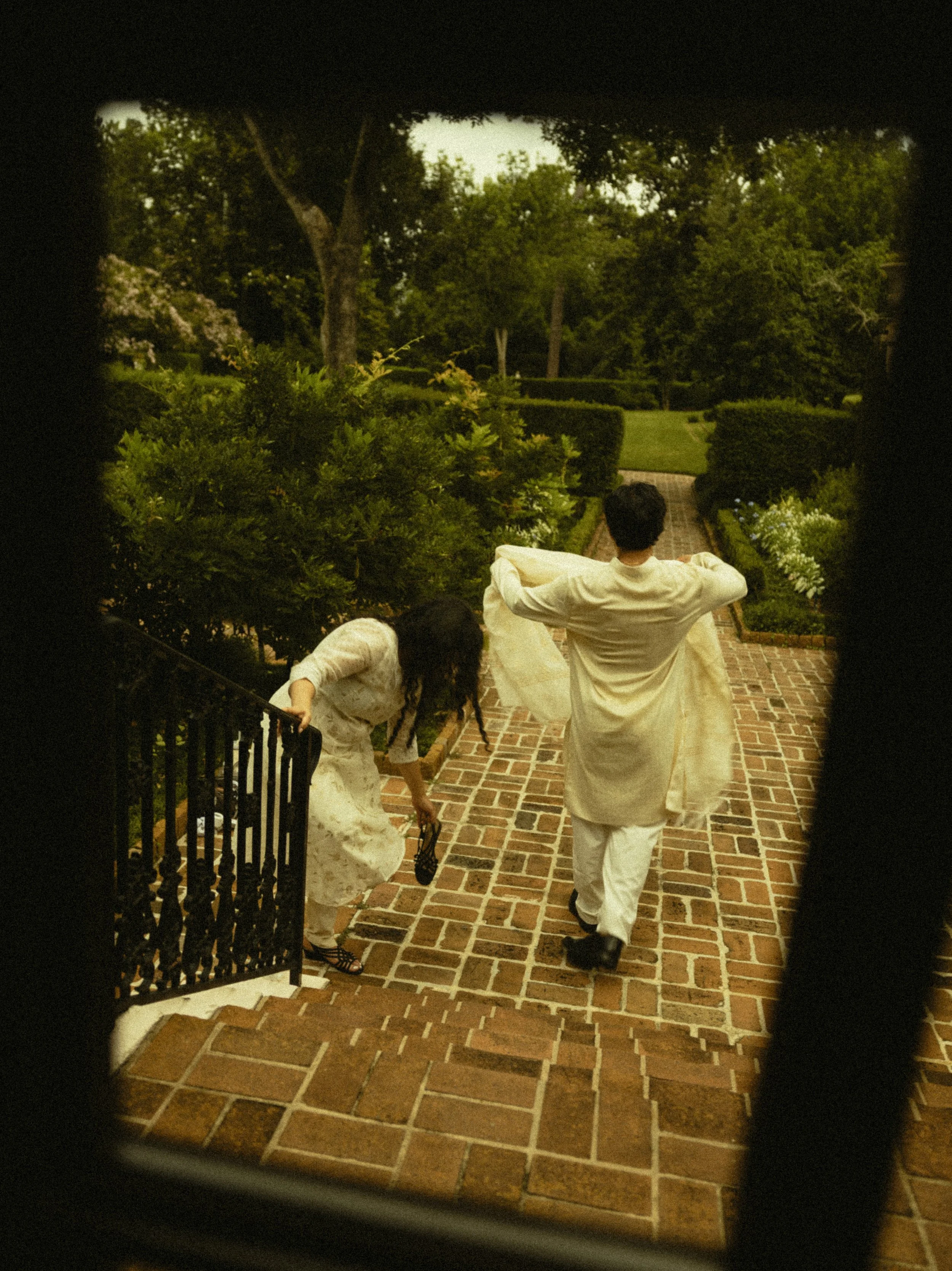 People walking down brick stairs outside, surrounded by greenery, viewed through a narrow opening.