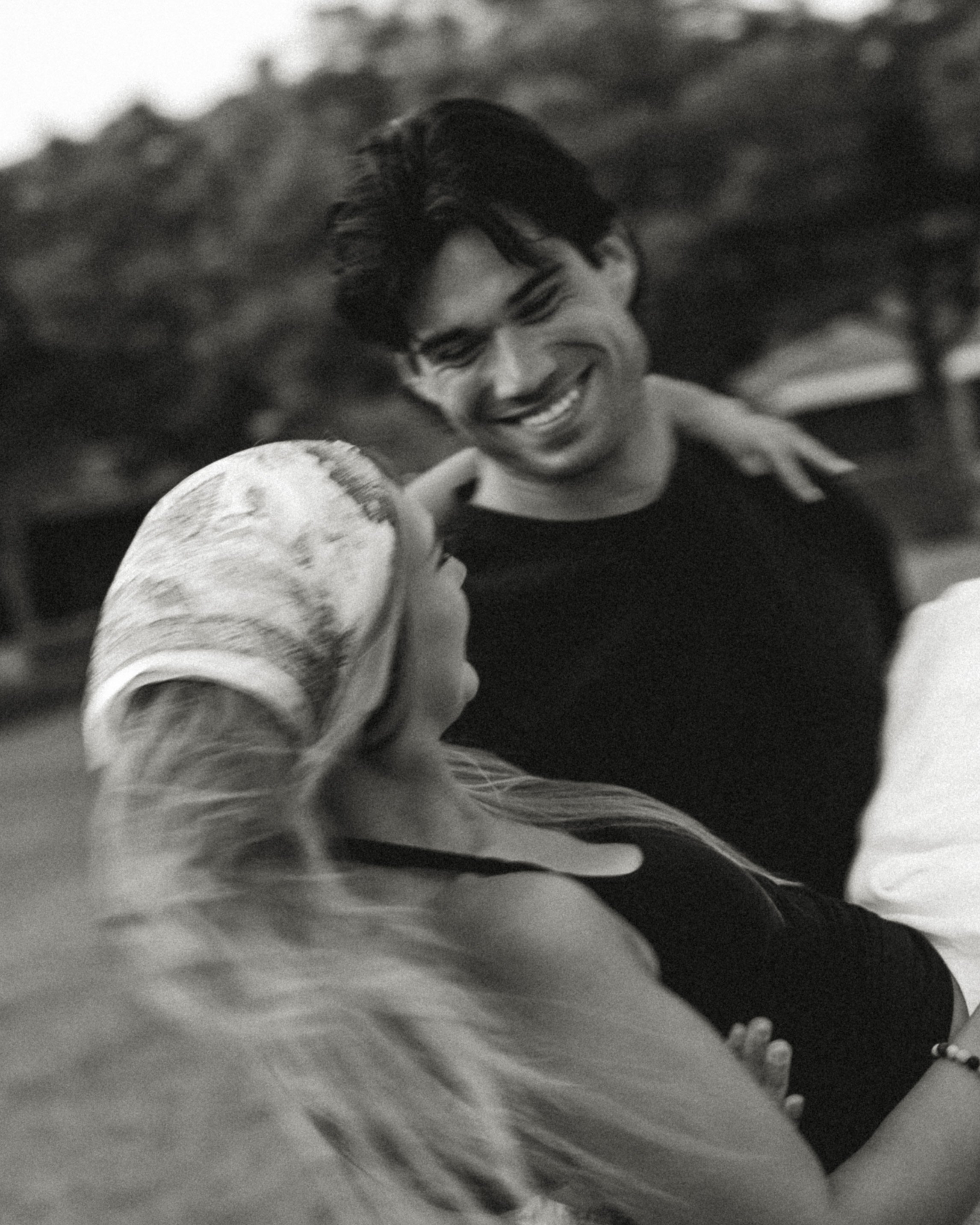 A young man with dark hair smiling and looking down, standing outdoors in a natural setting, wearing a black shirt and a necklace, with a woman wearing a floral dress.