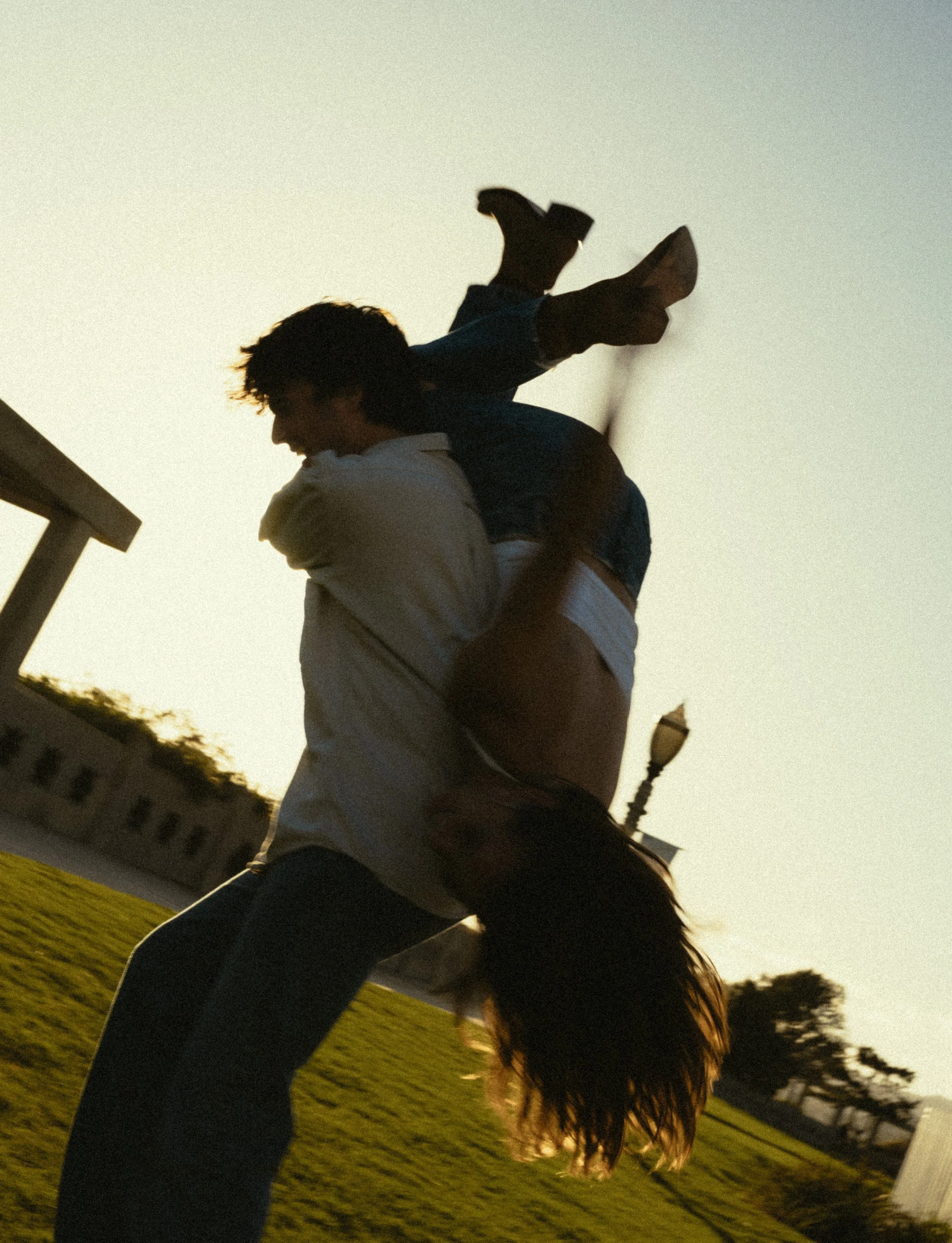 A person with long hair appears to be lifted onto another person's shoulders outdoors at sunset, with trees, buildings, and a streetlight visible in the background.