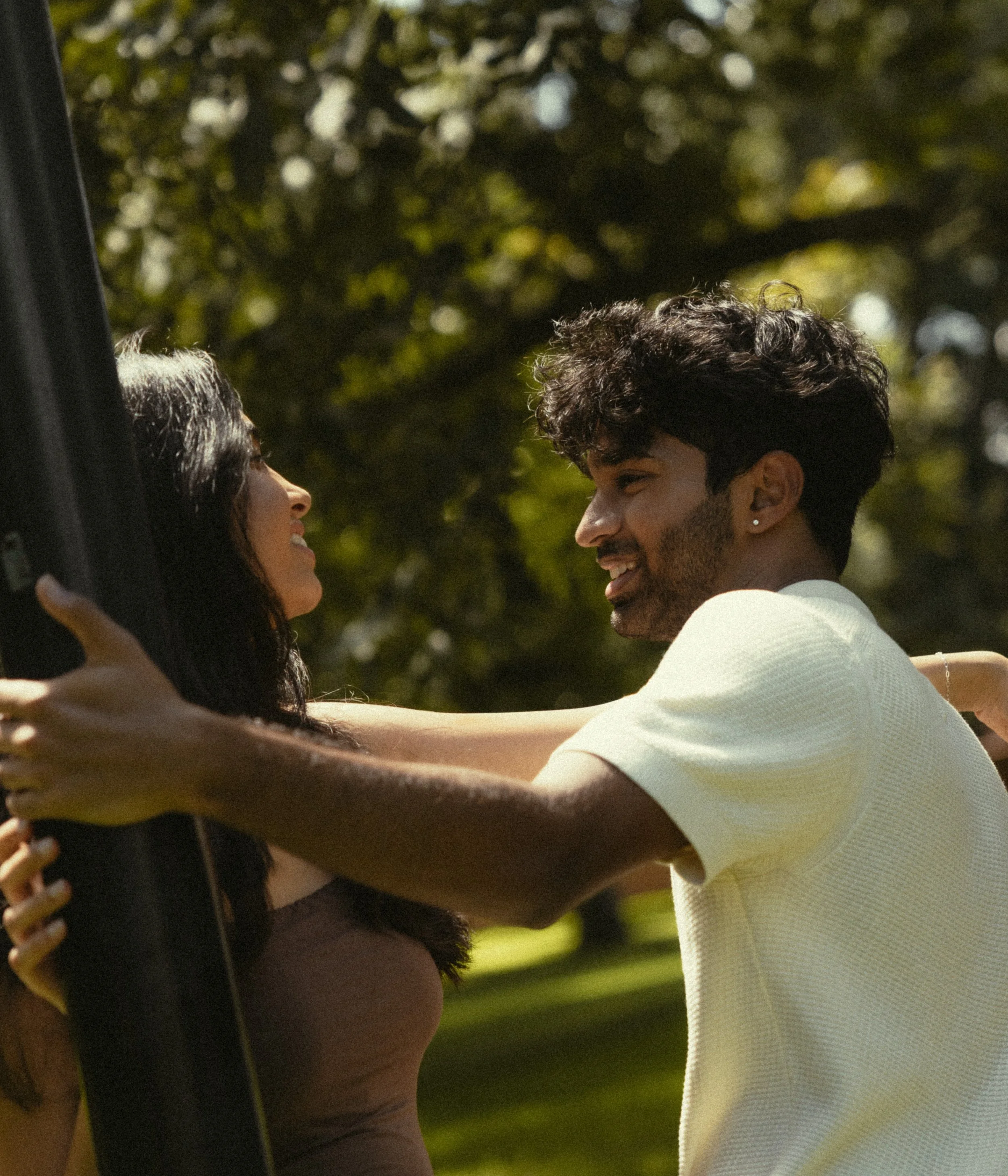 A young couple smiling at each other outdoors, with trees and sunlight in the background.