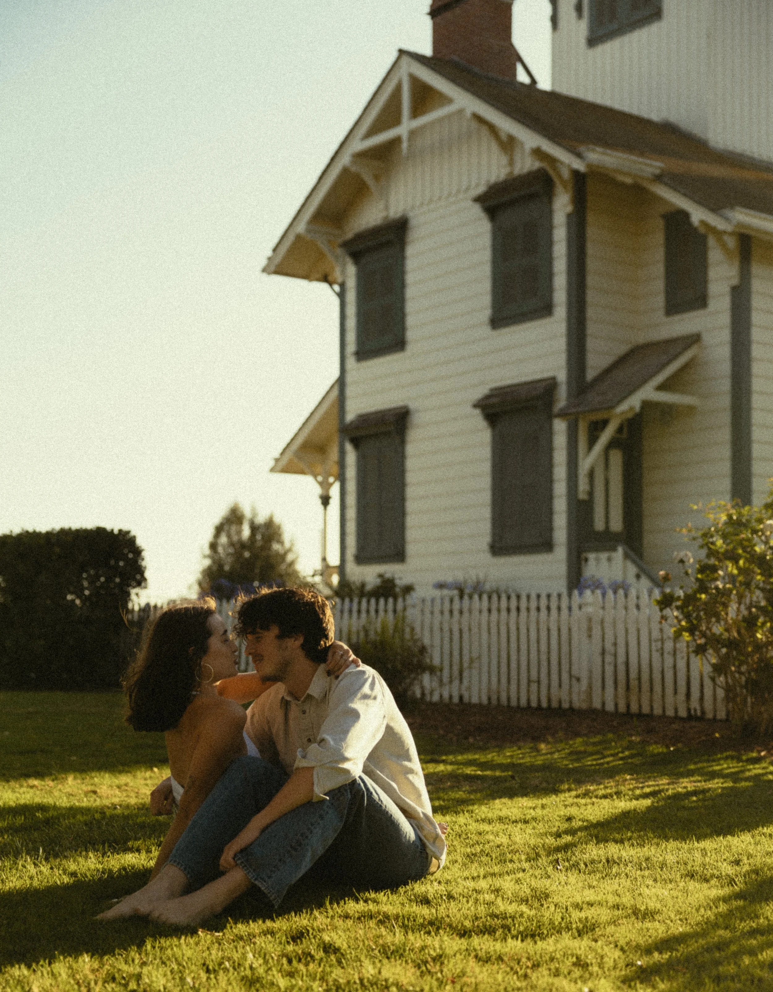 A young couple sits together on a grassy lawn outside a white house with dark window shutters at sunset, facing each other closely and smiling.
