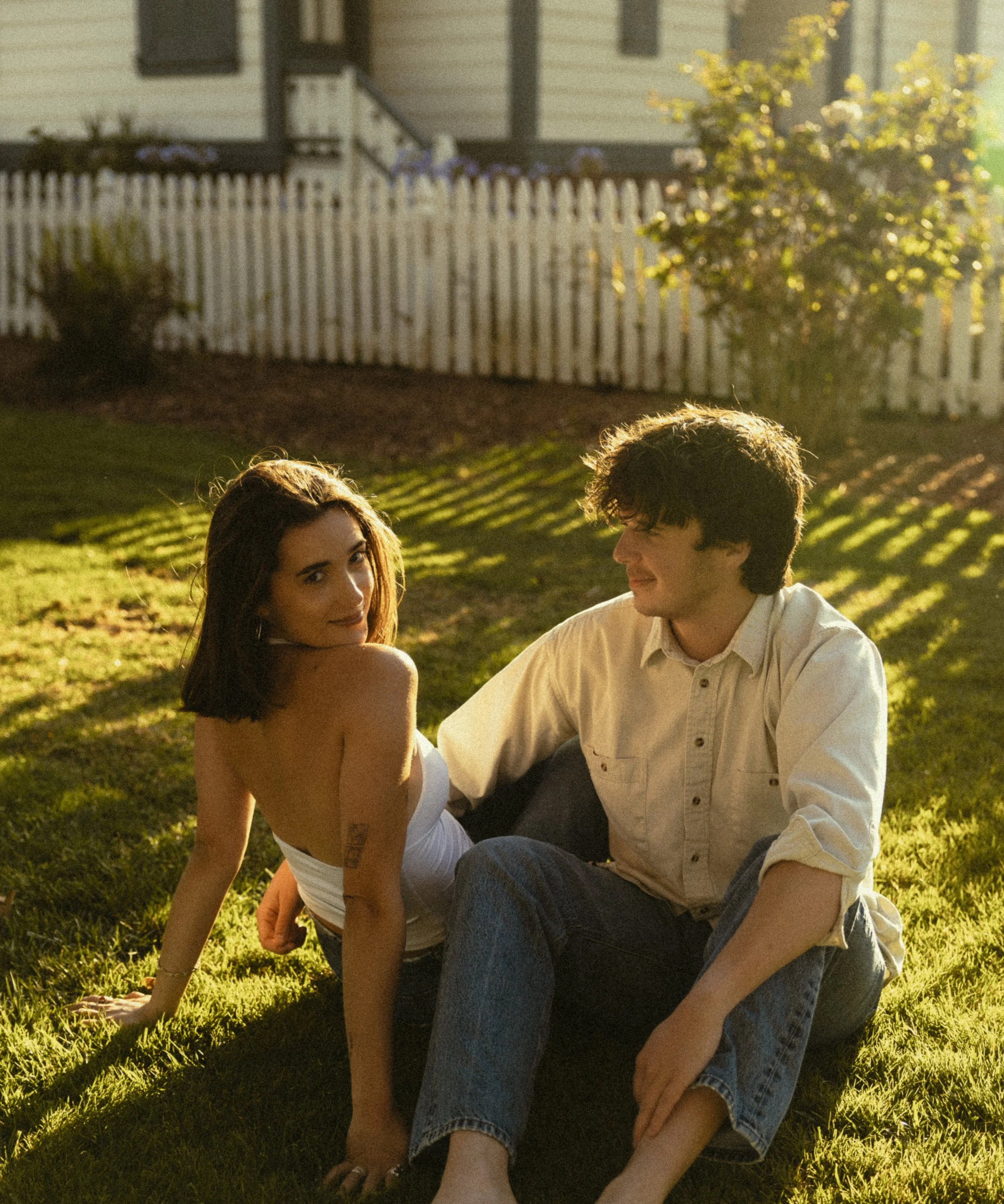 A young woman and a young man sitting on the grass in a backyard, facing each other, with a white picket fence, a house, and a tree in the background, lit by warm sunset light.