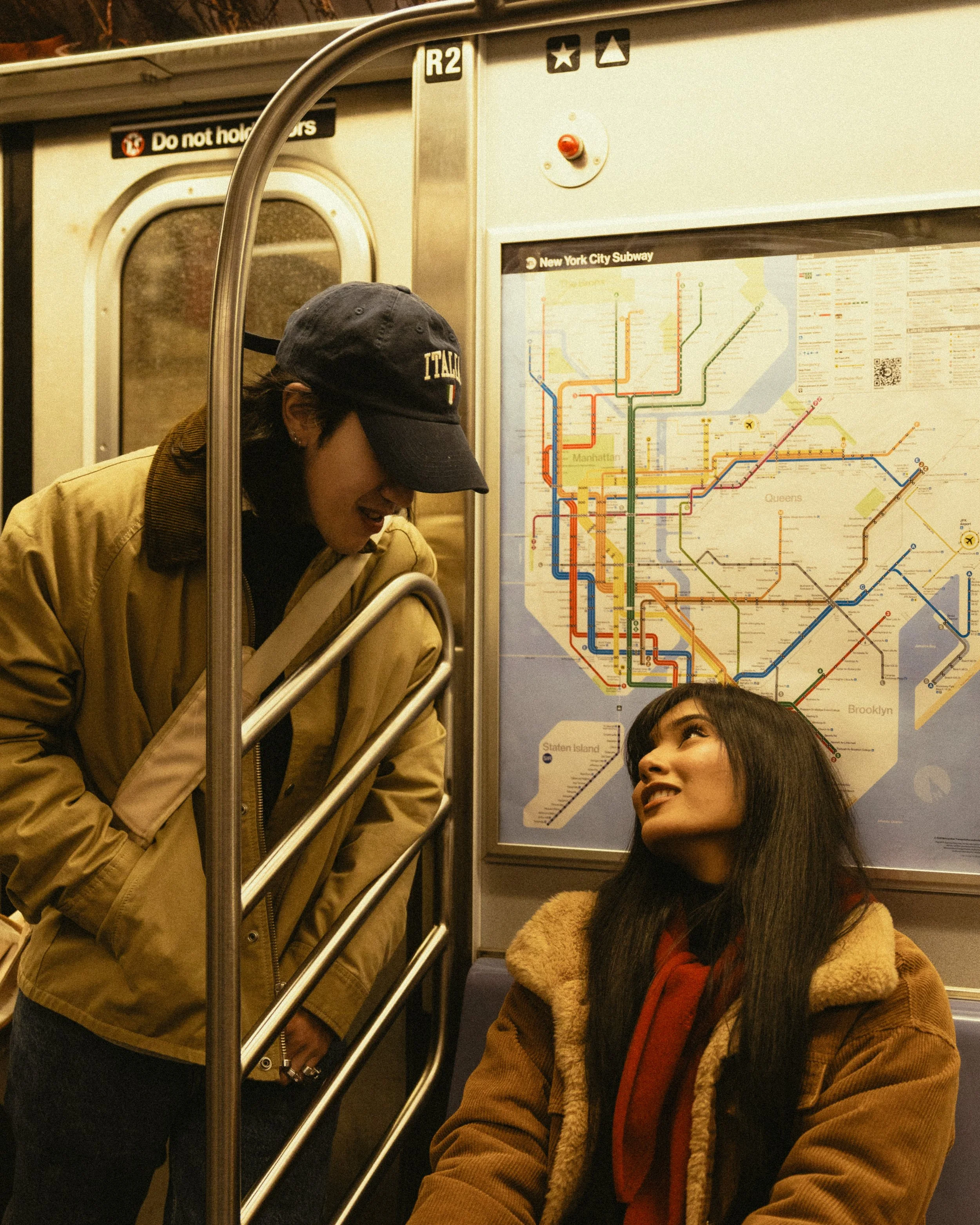 Two women on a New York City subway train. One woman with a black cap and tan jacket is leaning down and smiling at another woman with long dark hair and a brown coat, who is seated and looking up at her. A subway map is visible on the wall behind them.