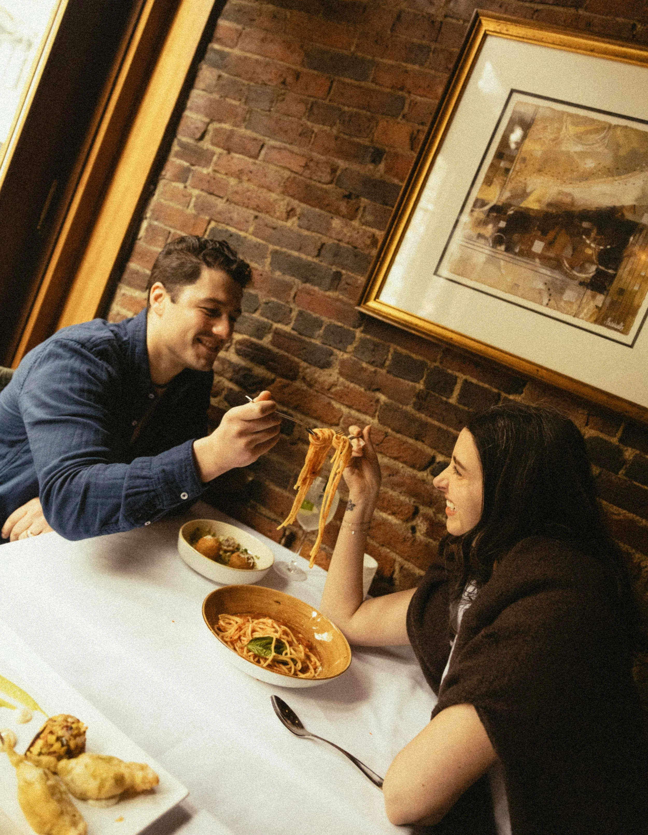 Two women and one man dining at a restaurant, sharing a plate of spaghetti. The man is smiling while holding a fork with a strand of pasta. The woman has dark hair and is smiling, holding a fork. The restaurant has a brick wall and framed artwork in the background. A white table holds bowls of pasta and fried appetizers.