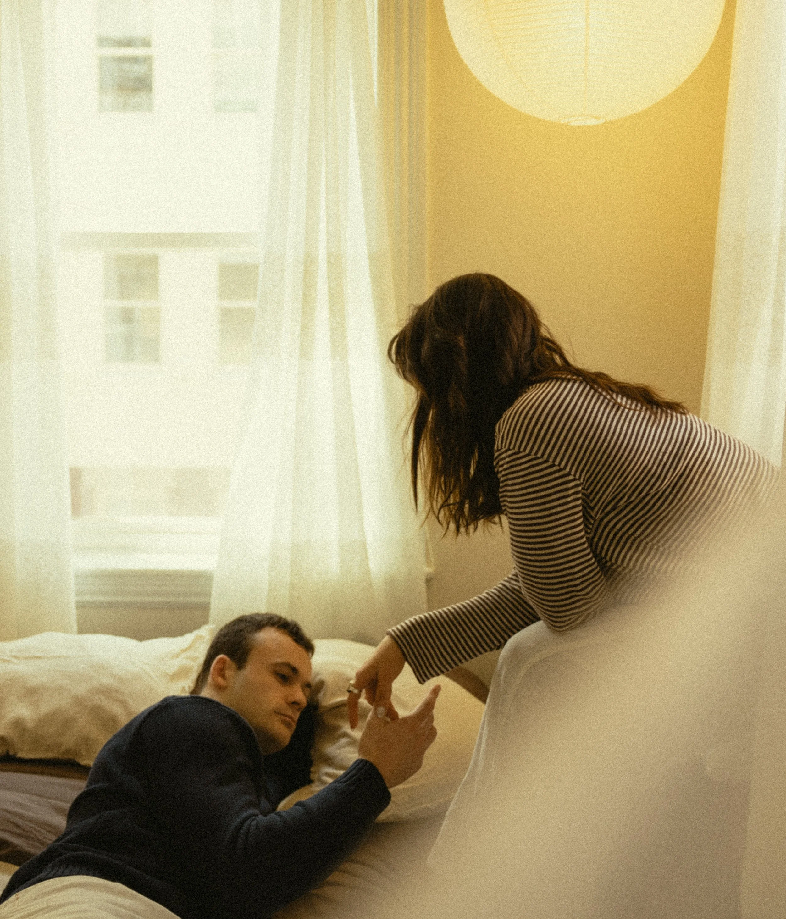 A woman with long curly hair wearing a striped shirt, kneeling on a bed, interacting with a man in a dark jacket lying down, in a room with cream-colored walls and a window with sheer curtains.