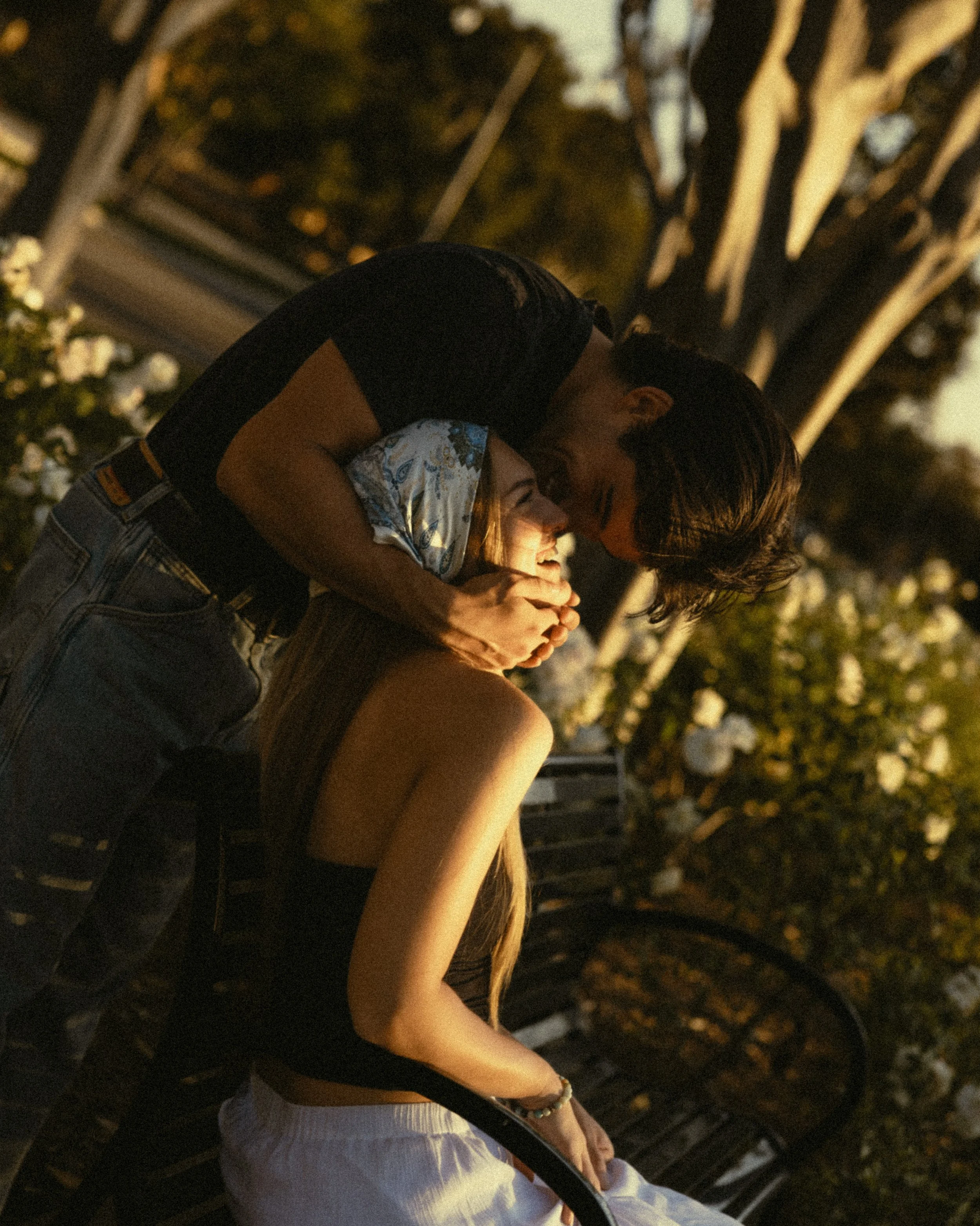 A young couple embracing and sharing a kiss outdoors during sunset, sitting on a park bench with trees in the background.