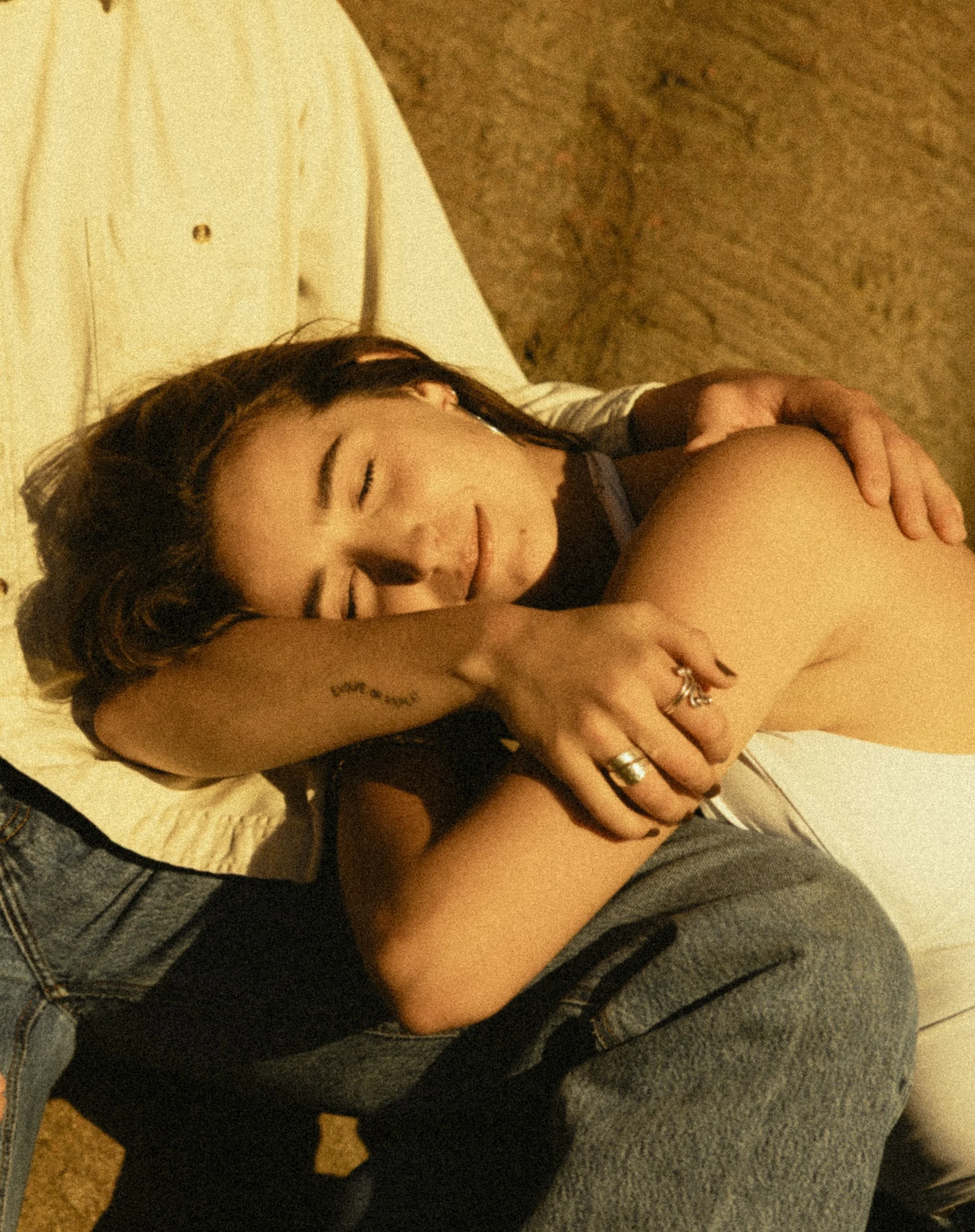 A young woman with dark hair and earrings, resting her head on a person's shoulder who is wearing jeans and a grey top, in a cozy indoor setting with a beige carpet and a cream-colored blanket.