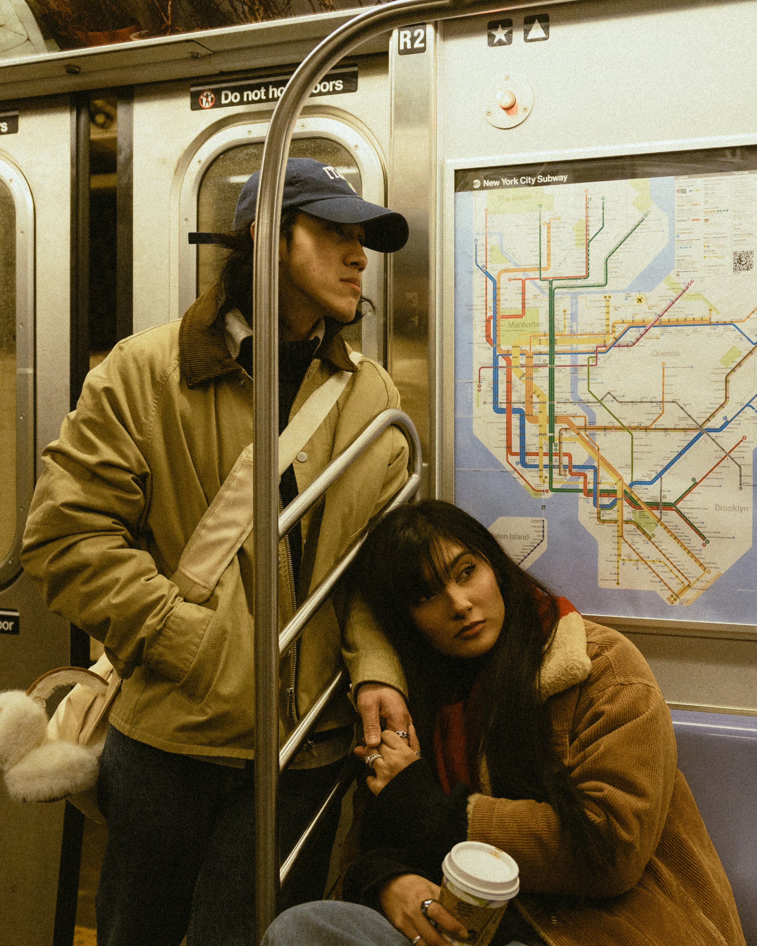 Two women sitting on the subway; one woman with long dark hair and tan coat, holding a coffee cup, leaning on the subway wall, and the other woman with dark hair, tan jacket, and red-checked shirt, resting her head on her hand with a serious expressi